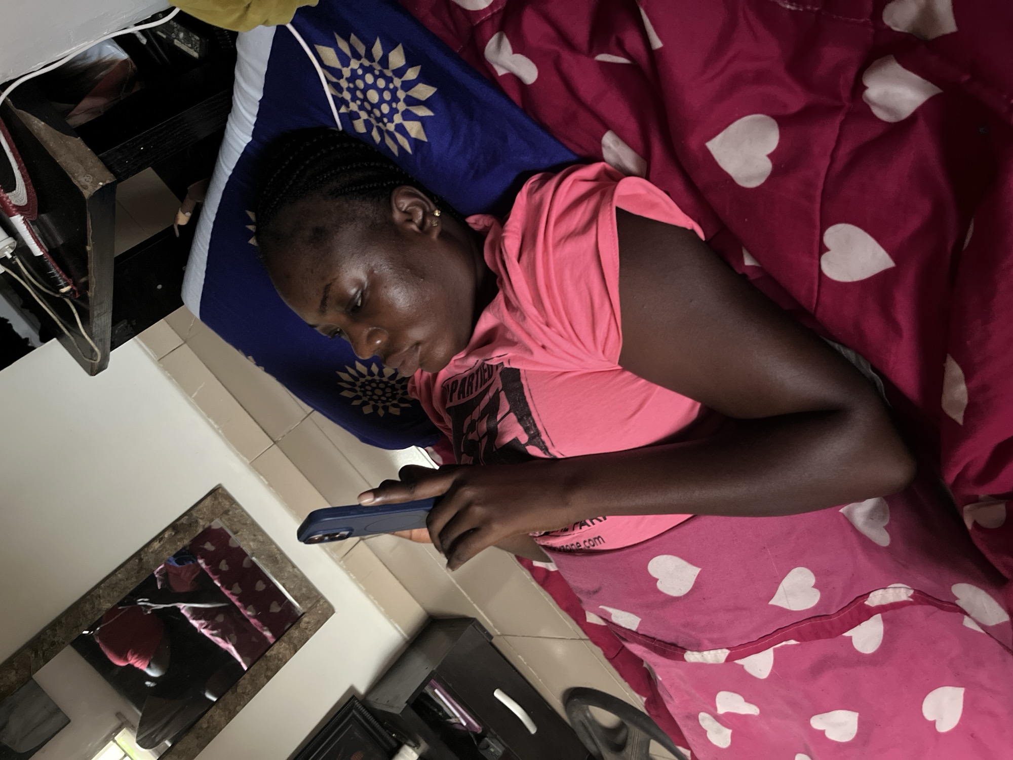 A young woman with dark skin and braided hair is depicted lying on a bed, engrossed in her smartphone. She is positioned on her left side, facing downwards towards the phone she holds in both hands. She wears a short-sleeved pink t-shirt, on which partial text "PARTIED" and what appears to be part of a web address like "one.com" are visible. A small gold earring is discernible in her right ear.
She is lying on a bed covered with a red/pink sheet or blanket adorned with white heart patterns. Her head rests on a blue pillow featuring a prominent gold or white circular, starburst-like pattern.
To the left of the bed, near her head, is a dark wooden or metal side table, where some items and white electrical cables are visible. The room has light-colored tiled flooring. A rectangular mirror with a patterned frame is positioned on the wall, reflecting parts of the room, including a dark cabinet or chest of drawers with a white handle, and a distorted reflection of the person on the bed. The overall impression is one of a casual, private indoor setting, likely a bedroom in Abuja, Nigeria, with the individual focused on their device.