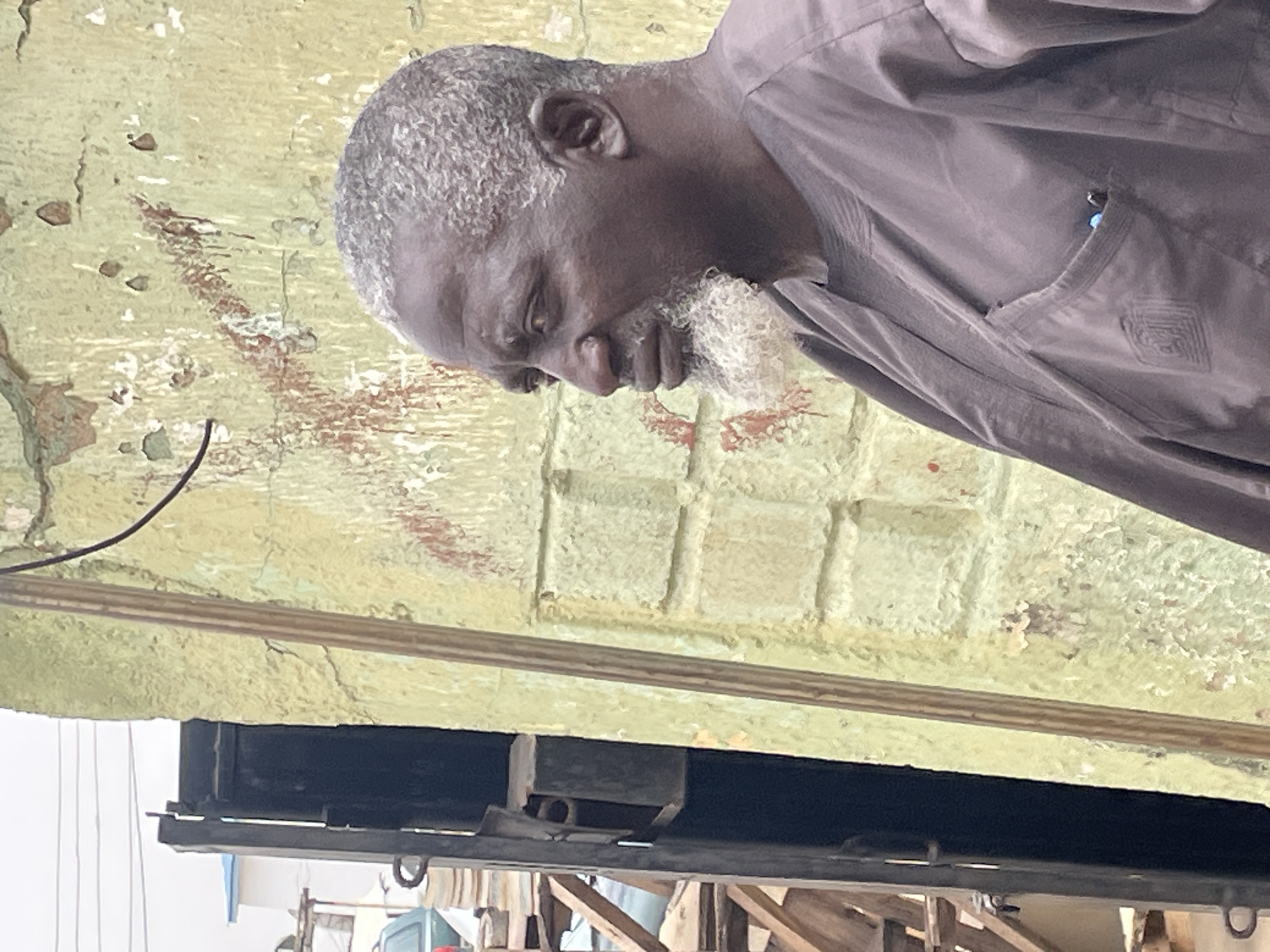 An older Black man with gray hair and a white beard looks down towards his right. He wears a brown button-down shirt. The man stands in front of a wall painted a greenish-beige color with brown markings. Below the wall, there is a black metal frame with hooks. The scene is in Jos, Nigeria.
