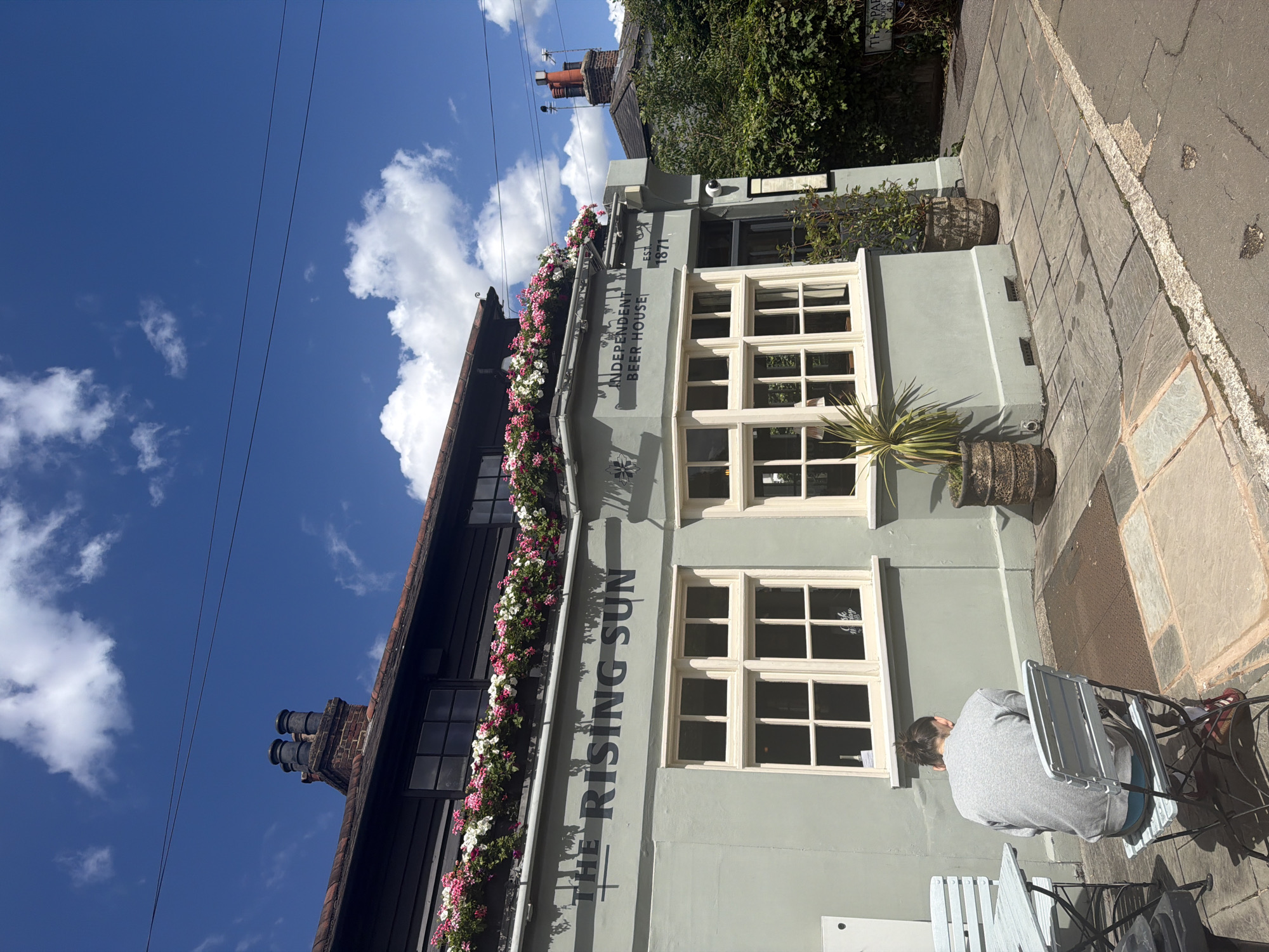 The image shows the exterior of "The Rising Sun," an independent beer house located in Epsom and Ewell, United Kingdom. The building is painted a pale green and features traditional windows with white frames. Flower boxes filled with pink and white flowers line the upper part of the building. The sign above the windows reads "THE RISING SUN" in large, dark lettering, with "INDEPENDENT BEER HOUSE" and "EST. 1871" also visible.
In the foreground, a person wearing a grey hoodie and blue shorts is sitting on a chair at a table outside the establishment. They are facing away from the camera, and their back is to the building. The scene appears to be set on a sunny day, indicated by the bright blue sky with white clouds and the strong shadows cast on the pavement. The pavement is paved with large, grey flagstones. A street runs alongside the pavement, with visible road markings. The overall atmosphere is relaxed and casual, suggesting a pleasant outdoor seating area at a pub.