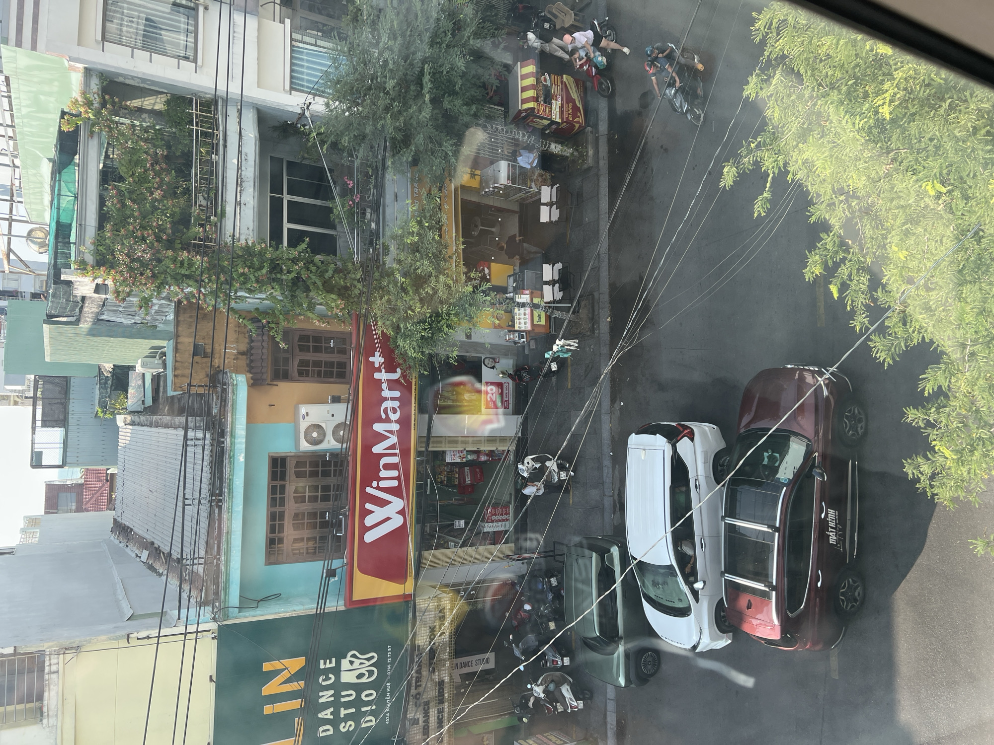 The image provides an overhead, high-angle view of a vibrant urban street scene, likely in Quy Nhon Ward, Vietnam, given the context. The perspective looks down onto the street, which runs diagonally from the bottom-left to the top-right of the frame, with buildings primarily lining the left side.
Several multi-story buildings are visible, showcasing a mix of residential and commercial establishments. One building is particularly green, covered in abundant foliage and flowering plants. At street level, a prominent red and yellow sign for "WinMart+" is clearly visible. Directly below it, a green sign identifies "LIN DANCE STUDIO" with the address "403A NGUYỄN HUỆ" and phone number "0766 72 73 57". Other businesses include small cafes or eateries with outdoor seating and a food cart marked "Bánh mì". Smaller signs with text like "20" and "SIÊU SỐC" are also present near WinMart+.
The street is bustling with activity. Numerous motorbikes, some carrying passengers, are either parked or in motion. Two cars are prominently parked near the right side of the street: a white SUV and a dark red SUV. The red SUV has "MẮT HÍTNH" written on its side. Several people are visible, either riding motorbikes, standing, or moving on the sidewalks. Overhead power lines crisscross the scene, a common feature in many Southeast Asian cities. The bright sunlight and clear shadows indicate it is daytime with fair weather. Lush green trees are interspersed throughout the urban landscape, adding natural elements to the concrete environment.