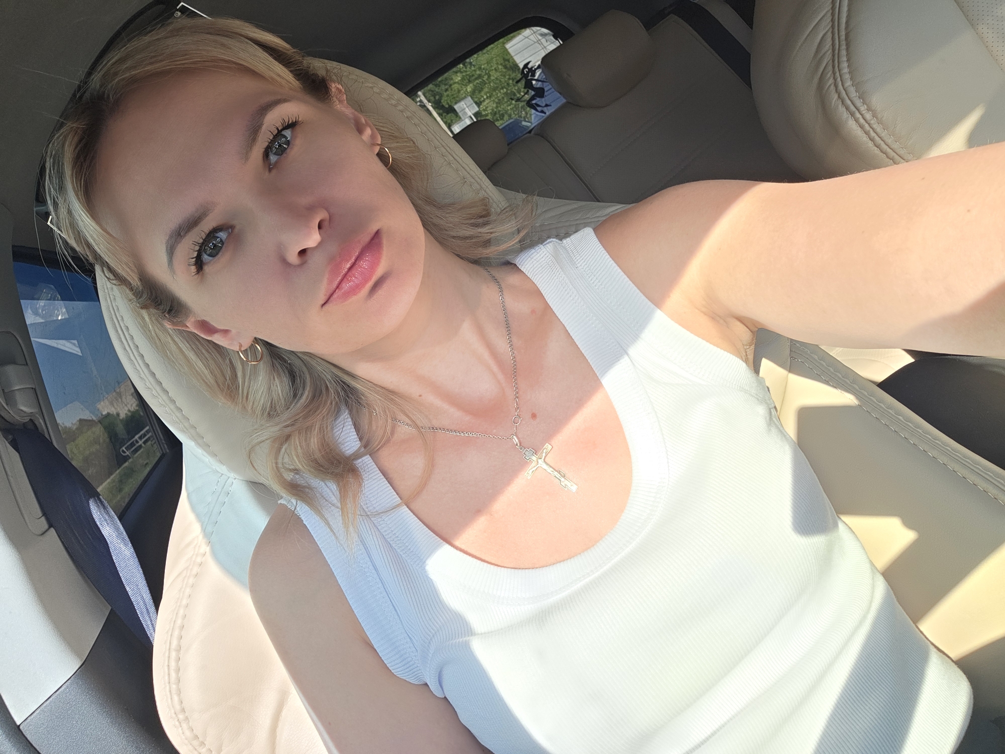 A woman is in the driver's seat of a car, looking towards the camera. She has blond hair and is wearing a white tank top and a necklace with a cross pendant. The car has light-colored leather seats. Through the window, there is a glimpse of greenery and a vehicle outside, possibly in the city of Chaikovsky, Russia.