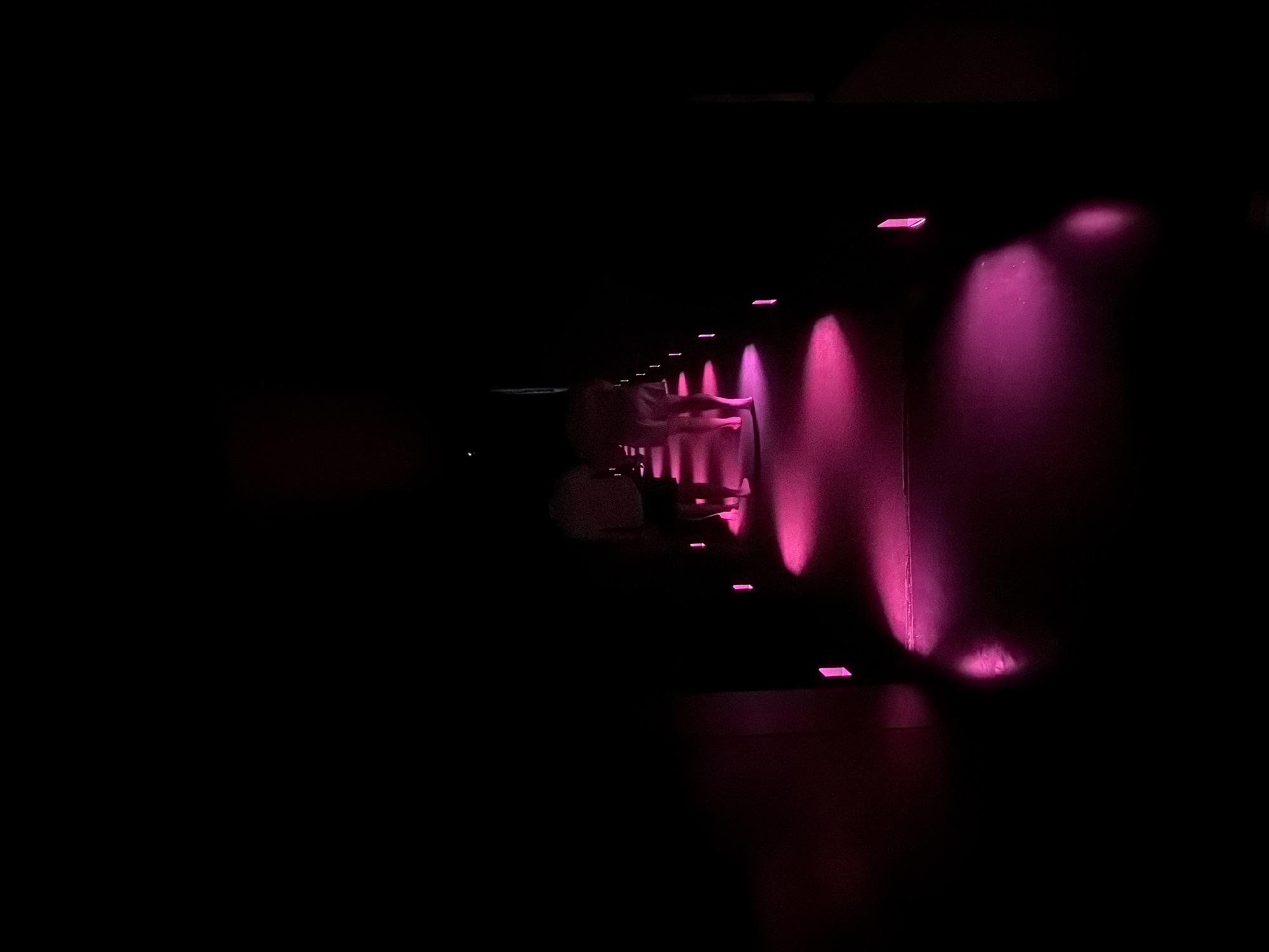 The image shows two individuals from behind, walking down a dimly lit corridor or pathway. The scene is illuminated by numerous pink lights embedded in the ground, casting a vibrant glow on the floor and the lower parts of the walls. The individuals appear to be wearing casual clothing, with one wearing shorts and the other with darker lower garments. The overall atmosphere is dark and somewhat mysterious, with the pink lighting creating a stark contrast against the blackness of the surroundings. This scene could be an art installation or a specially lit pathway within a venue. The location in Koto, Japan, is not discernible from the visual information provided in the image.