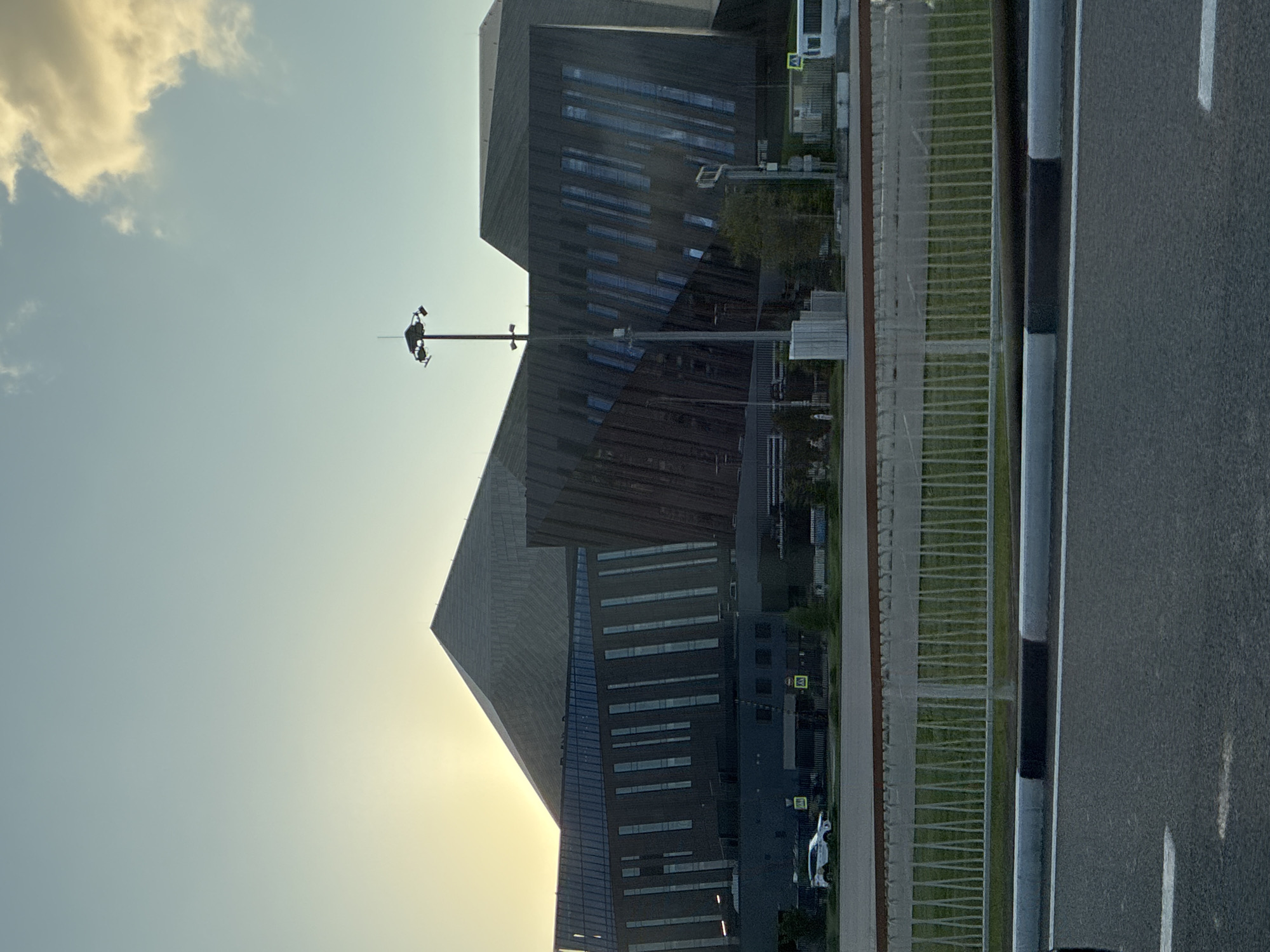 The image captures a contemporary building complex with striking geometric architecture, characteristic of the Sirius federal territory in Russia. The structures feature a combination of dark, angular facades and large, vertical windows. A prominent feature is a tall, multi-armed lighting mast situated in front of the buildings. In the foreground, a paved road with white and black markings is visible, separated by a curb from a grassy area and a white metal fence. A white vehicle is parked on the road behind the fence, and a white bus is seen in the background. The sky above is a pale blue, with a few wispy clouds, and the sun is setting or rising, casting a soft, warm light. The overall scene suggests an urban environment with a focus on modern infrastructure and design.