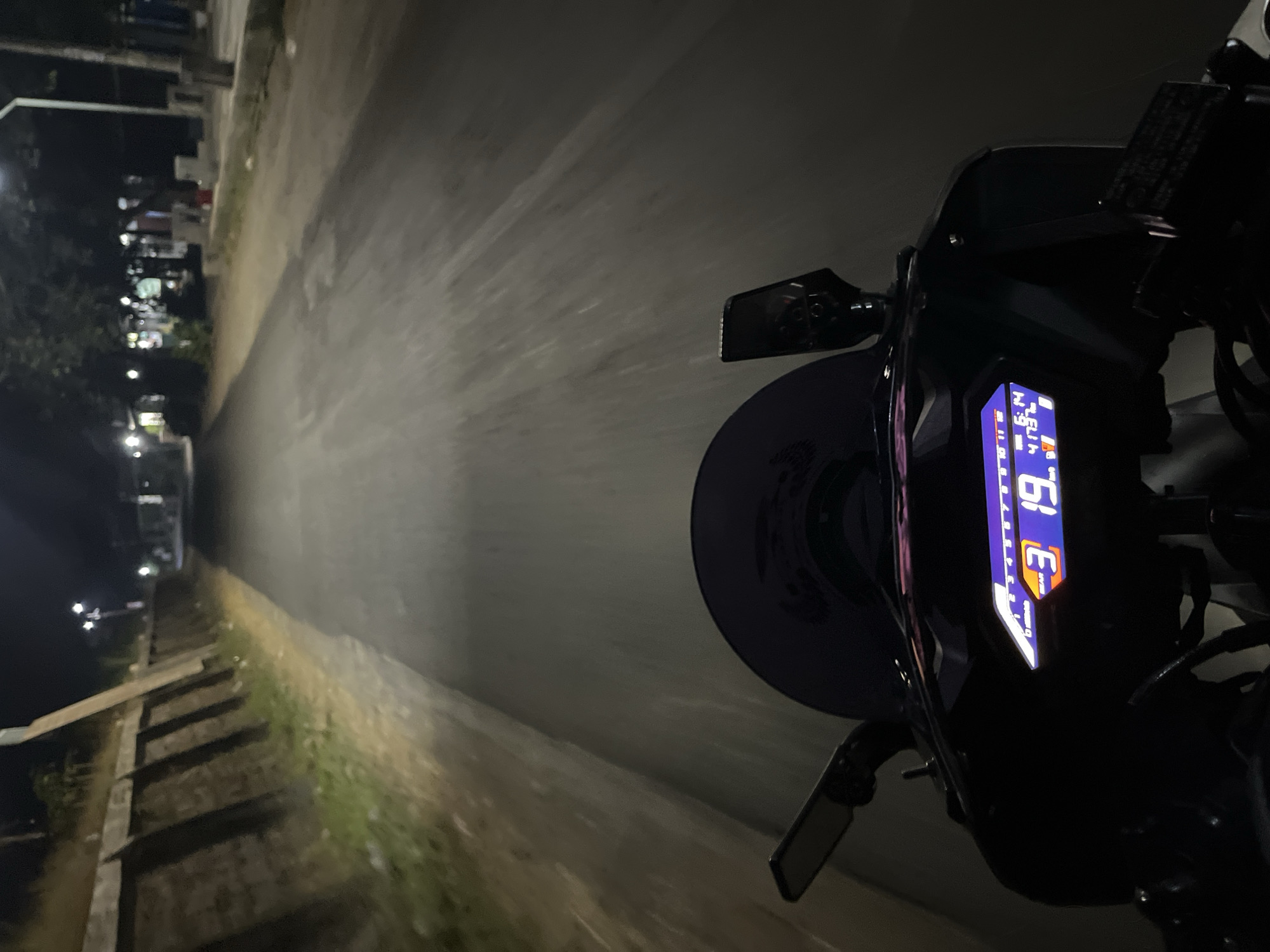 A first-person perspective video of someone riding a motorcycle at night through a moderately lit street. The road is paved and appears to be a two-lane street with cars parked on the right side. The motorcycle's digital dashboard displays a gear indication of "3", a speed of "19", a clock reading "6:14", and an odometer reading of "4173". On the left side of the road, there is a raised concrete barrier with vegetation growing on top. On the right side of the road, there are buildings and streetlights, suggesting an urban or semi-urban environment. The overall lighting suggests it is nighttime. The video captures a sense of movement as the road ahead is blurred. The location is identified as Karangrandu, Indonesia.