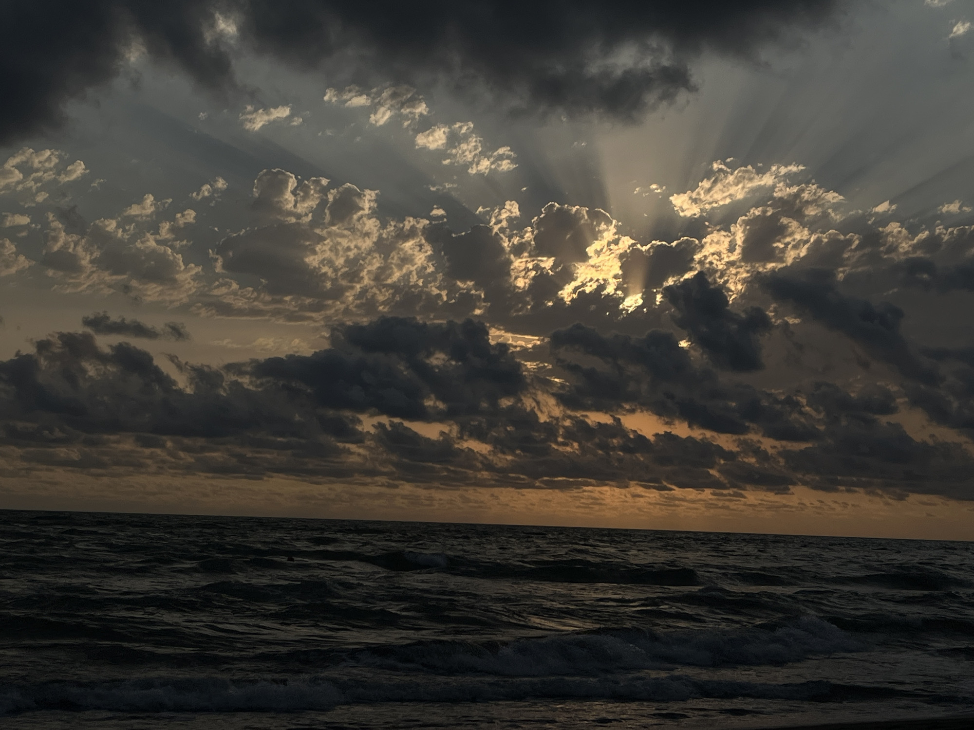 The image shows a dramatic sky over a dark, choppy sea. The sun is setting or rising, casting crepuscular rays through breaks in dark, heavy clouds. The light filters through the clouds, creating a striking contrast between illuminated and shadowed areas of the sky. The sea below is a deep, dark color, with visible waves and whitecaps. The horizon line is clearly defined, separating the turbulent water from the dynamic sky. The scene appears to be a coastal area, possibly the Black Sea coast near Supsa, Georgia, given the presence of such dramatic weather conditions. There are no people or man-made objects visible in the frame, emphasizing the raw power of nature.