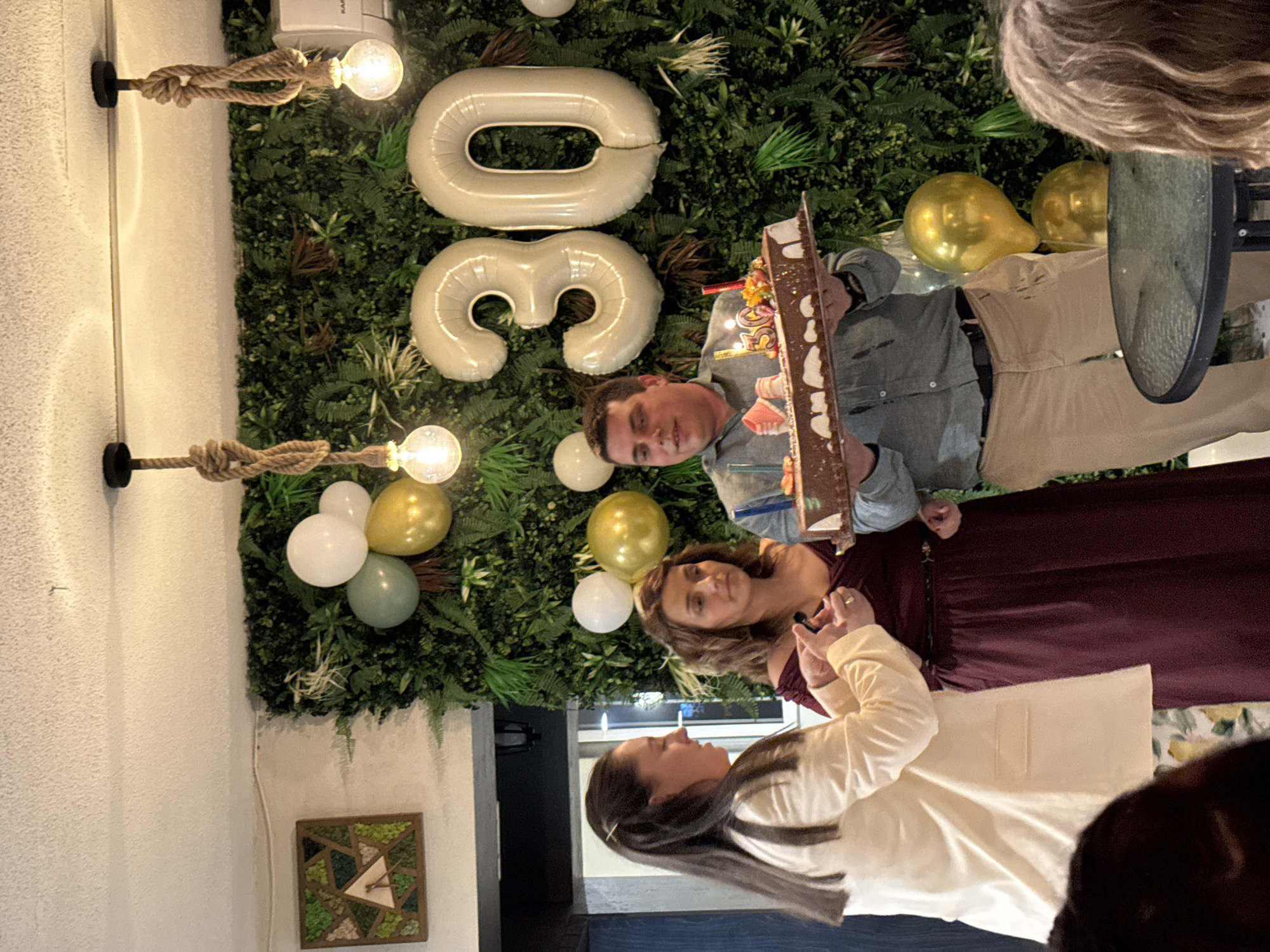 The image depicts an indoor event celebrating a 30th birthday, suggested by the number "30" in large white balloons. A man holds a decorated cake with lit candles, while two women stand nearby. The background features a wall decorated with greenery, balloons, and wall-mounted light fixtures. A small, framed geometric artwork is visible on the wall. Based solely on the information within the image, there is no way to determine if this event happened in the city of Buhovo, Bulgaria.