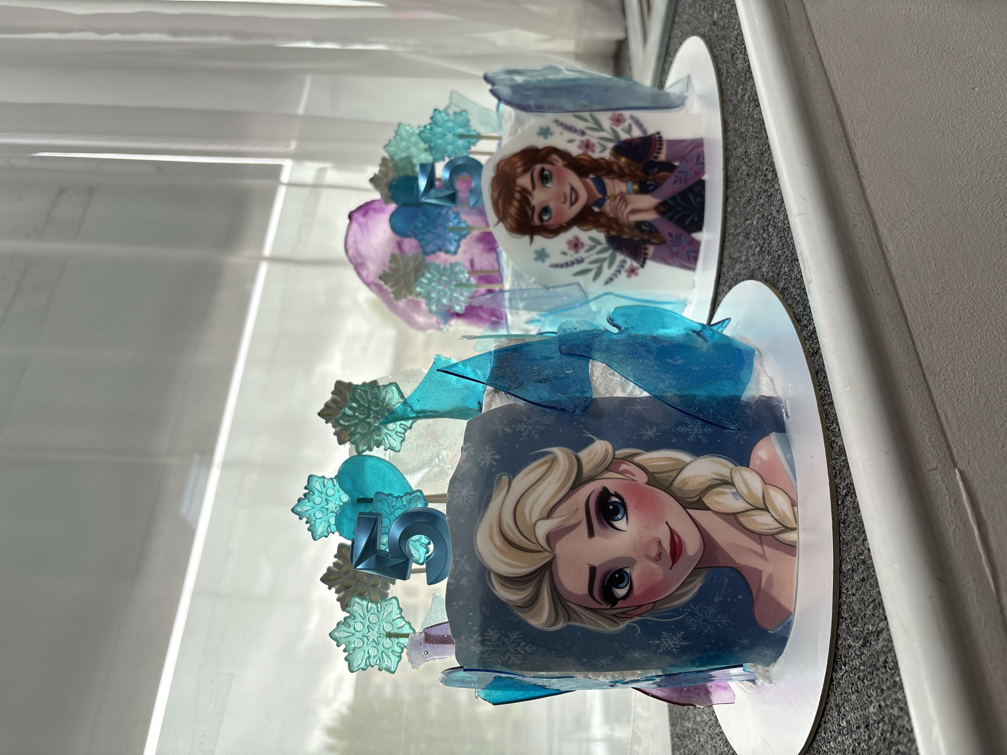 Two cakes decorated with images from the Disney film "Frozen" are displayed. The cake on the right features Elsa, while the cake on the left features Anna. Both cakes are adorned with candy shards and snowflake decorations. A number "19" is on top of the Elsa cake. The cakes are positioned near a window with blinds. The exact location in Krasnoyarsk, Russia, is not discernible from the image.