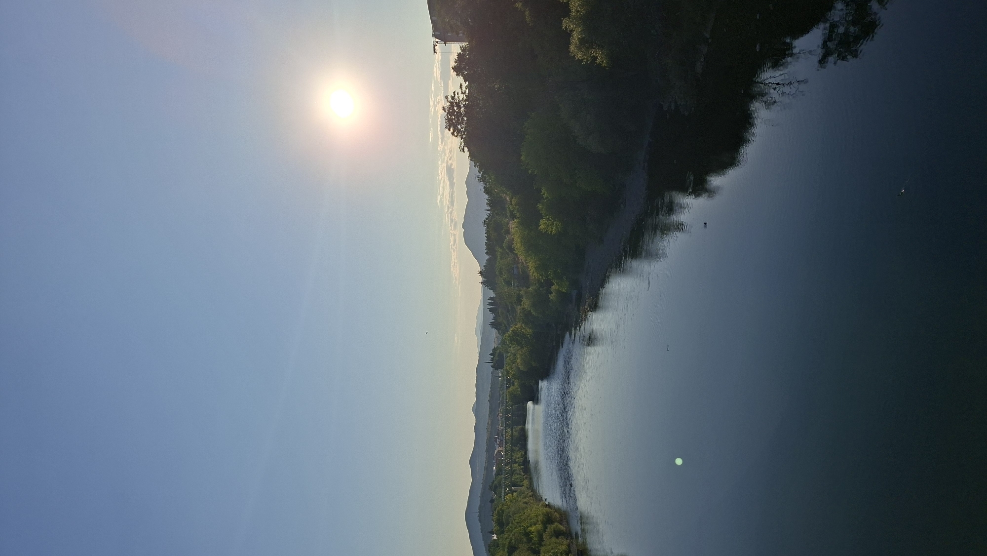 The image depicts a wide river flowing through a verdant landscape under a clear blue sky with the sun shining brightly. The river's surface is calm, reflecting the sky and surrounding trees. On the left bank, lush green trees line the shore, leading to rolling hills and mountains in the distance. On the right bank, more trees and some structures are visible, including a tall building or crane. A bridge can be seen spanning the river in the middle ground, with buildings and more trees on either side. The scene appears to be a serene natural setting within or on the outskirts of a city. This is a view of the Morača River in Podgorica, Montenegro. There are no people or specific actions depicted in the image.