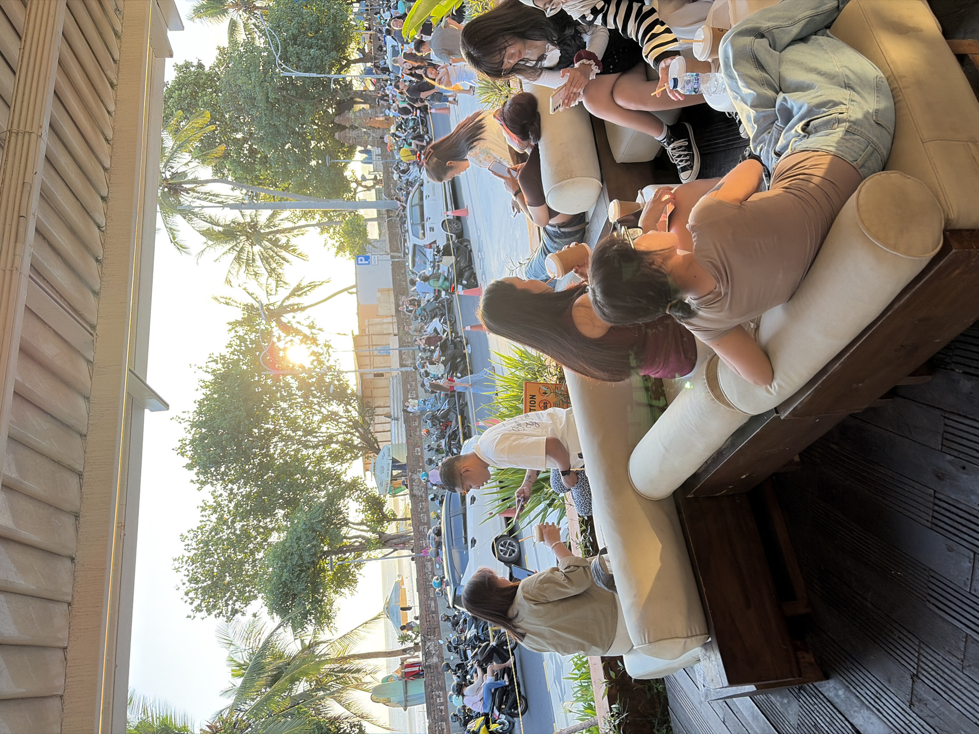 The image depicts a lively street scene in Kuta, Indonesia, likely near the beach, as indicated by the presence of palm trees and surfboards visible in the background. The foreground features a group of people relaxing on outdoor seating, possibly at a cafe or restaurant. The setting sun casts a warm glow over the scene, with palm trees silhouetted against the bright sky.
In the immediate foreground, several individuals are seated on large, light-colored sofas, engaged in conversation or using their phones. One woman in a light brown shirt is holding a drink and looking at her phone. A man in a white t-shirt is seated next to her, also looking at his phone. Further back on the sofas, other people are visible, some with drinks in hand.
Beyond the seating area, a street is bustling with activity. Numerous scooters and motorcycles are parked along the roadside, suggesting a popular area for transportation. Cars are also present, navigating the street. People are walking along the sidewalk and crossing the street. In the background, the ocean is visible, with gentle waves. The overall atmosphere appears relaxed and social, characteristic of a seaside town in Indonesia during sunset.