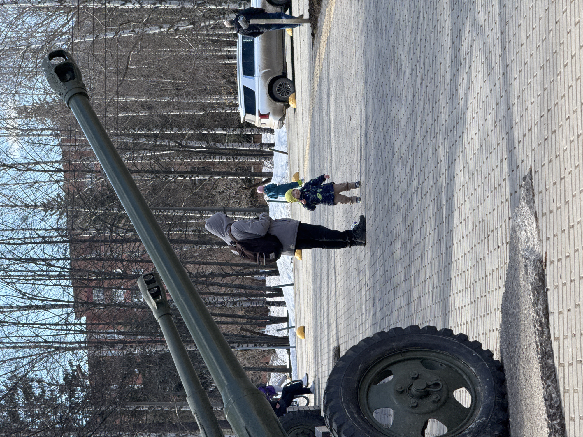 A medium outdoor shot shows a museum or park exhibit of two antique military cannons alongside a paved walkway. One cannon's barrel rises diagonally toward the upper left, while its tire is placed in the lower right of the shot. A second cannon is partially visible. A pedestrian in a gray hooded jacket is walking across the sidewalk, and a toddler in a bright yellow hat and patterned blue coat is near them. In the background, bare birch trees are visible. A white car is parked behind the trees. This is a common sight in Izhevsk, Russia where such monuments and military exhibits are publicly displayed.