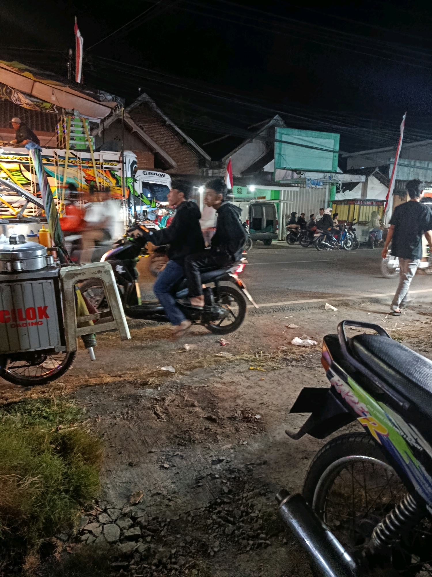The image depicts a nighttime scene in what is identified as the city of Kedok, Indonesia. In the foreground, a motorcycle is partially visible, with its rear wheel and exhaust pipe in focus. To the left, a mobile food cart, labeled "CILOK DAGING," is positioned next to a small plastic stool. In the mid-ground, two young men are riding on a motorcycle, with the rider wearing a black hoodie and blue jeans, and the passenger wearing a black hoodie and black pants. They are moving away from the viewer. Behind them, several other motorcycles are parked or in motion, with other individuals visible in the background. Structures, possibly market stalls or shops, are visible along the street, some adorned with Indonesian flags. The environment appears to be a street or roadside area, with a paved surface and some patchy grass and debris. The lighting suggests artificial illumination from streetlights or nearby businesses.