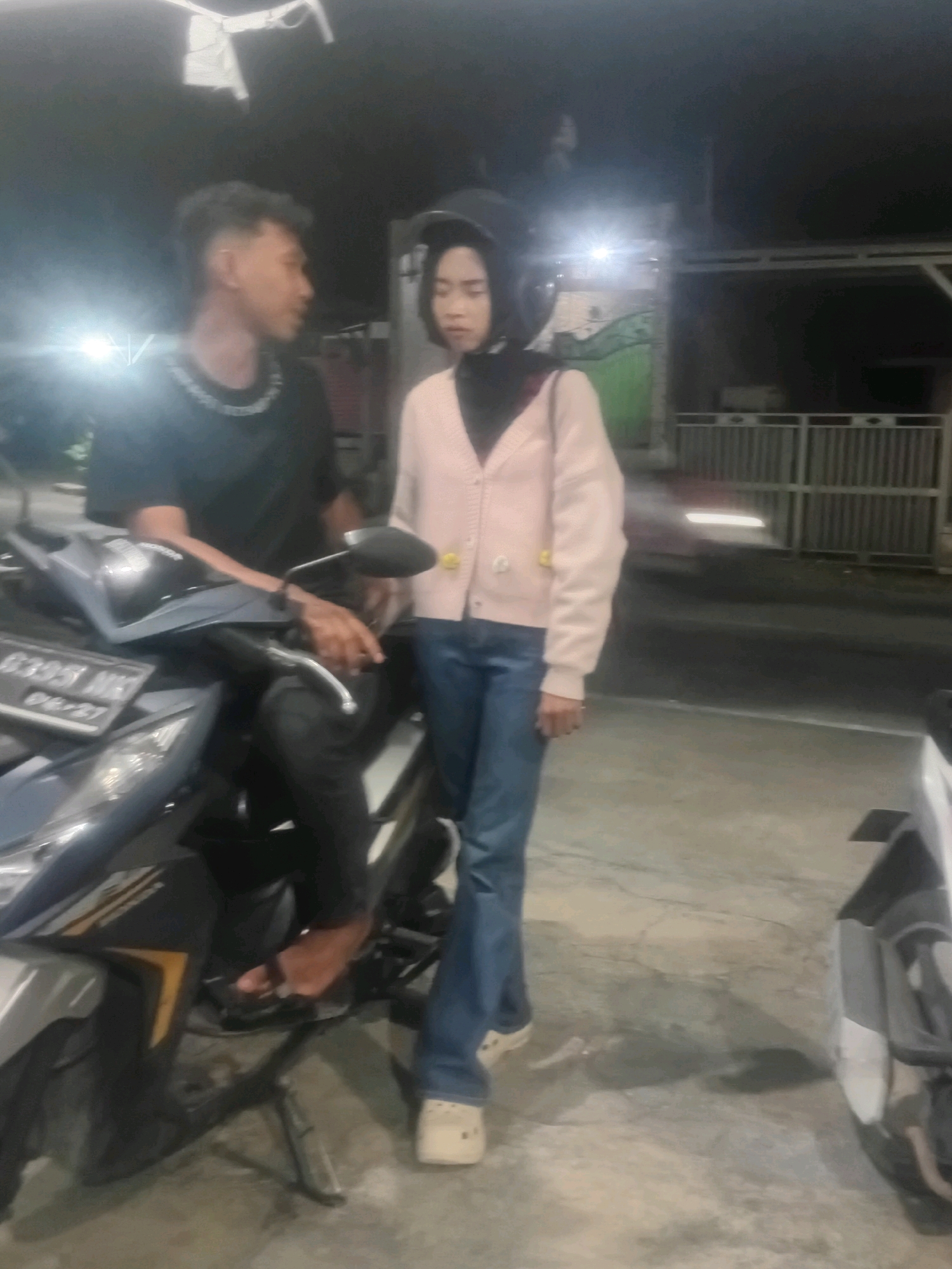 A medium-shot image captured at night shows a young man and woman standing next to a motorcycle on a paved surface, likely a street or parking lot in Bae, Indonesia. The man, on the left, is seated on the motorcycle, wearing a black t-shirt with a silver chain detail and black pants. He has short, styled dark hair and is looking towards the woman. The woman, to the right, stands beside him, wearing a light pink cardigan over a dark hijab and blue jeans. She is also wearing a helmet and looking slightly downwards. The motorcycle, a modern scooter, is gray and black with visible license plate information. In the background, a metal fence is visible, along with a building and some greenery, all illuminated by artificial lighting, suggesting an urban or semi-urban setting in Bae, Indonesia. The overall scene appears casual, with the couple appearing to be in conversation or pausing during a journey.