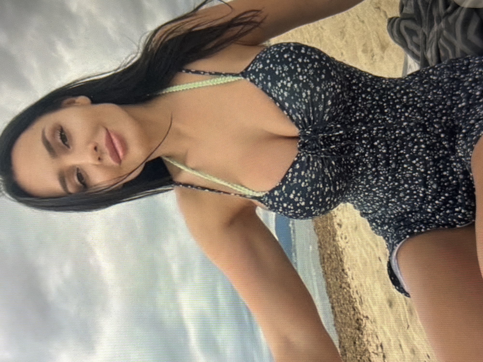 The image shows a young woman with long dark hair and fair skin sitting on a beach. She is wearing a dark blue floral print dress with spaghetti straps. The background consists of the beach, the sea, and a cloudy sky. There are no visible landmarks or elements that would suggest the location is in Shumen, Bulgaria.