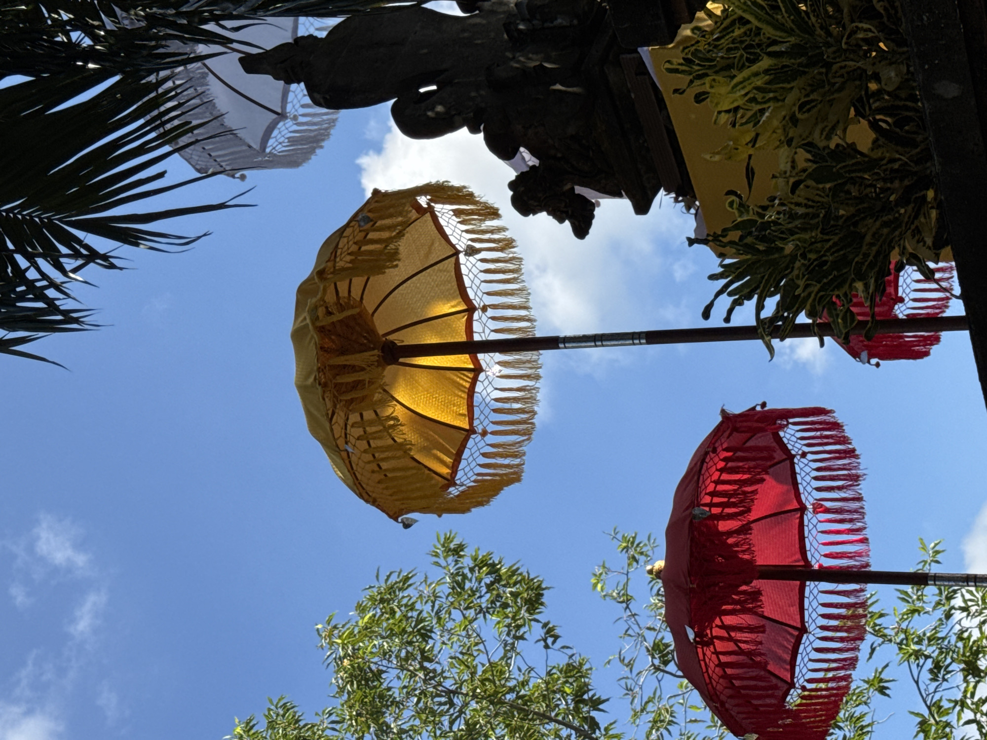 In the city of Ubud District, Indonesia, beneath a bright blue sky with scattered white clouds, several decorative umbrellas are visible. The most prominent are a large yellow umbrella with gold fringe and a smaller red umbrella with red fringe, both supported by dark poles. To the right, a dark stone statue or carving is partially obscured by a palm frond. In the background, a white umbrella with white fringe is also visible. Lush green foliage from trees and plants frames the scene. The image is captured from a low angle, looking upwards towards the sky and the decorative elements.