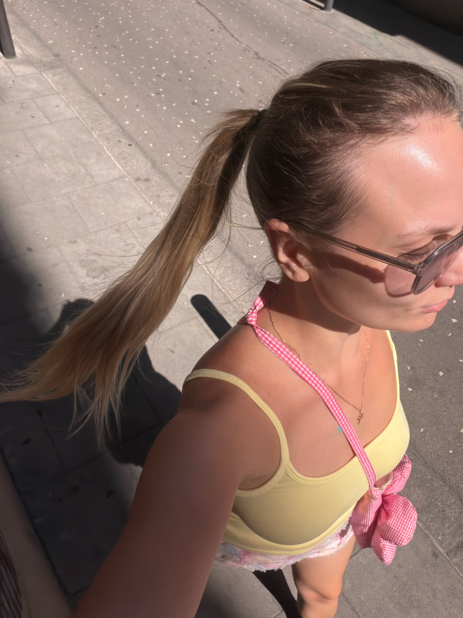This is a close-up, high-angle, outdoor shot of a young woman walking on a sidewalk in Barcelona, Spain, likely during the day. The woman, seen from the chest up and slightly from the side, has blonde hair pulled back in a ponytail, and is wearing sunglasses, a yellow tank top, and a pink gingham strap across her shoulder. A small pink gingham bag is slung over her shoulder. The sidewalk is paved with light-colored square tiles, and a shadow of the woman, indicating the sun's position, is cast on the ground to her left. The background is blurred, but appears to be a street with a slight incline.