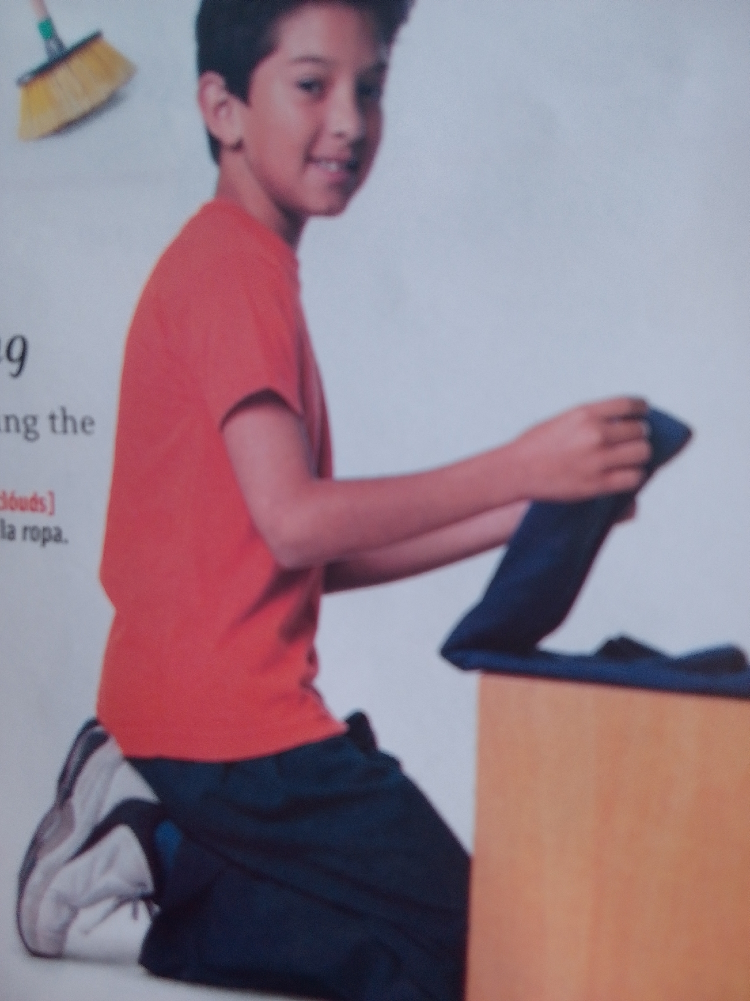 In the foreground, a young boy with dark hair and brown eyes kneels, facing left. He is wearing a bright orange t-shirt and dark blue pants. His left foot, clad in a white and grey sneaker, is visible. In his hands, he is holding and folding a dark blue garment, possibly pants or a shirt, which rests on a light brown surface, likely a table or box. The boy appears focused on the task.
To the upper left of the image, a yellow broom with a wooden handle is partially visible, suggesting a domestic or cleaning-related context.
The background is a plain, light color, indicating an indoor setting. There is some text visible on the left side of the image, though it is partially cut off and blurry. The visible text includes "9", "ng the", and "la ropa.". Based on the context of the image and the visible text, it's plausible this is a page from a children's book or educational material related to chores or daily activities. The overall impression is one of everyday life and learning.