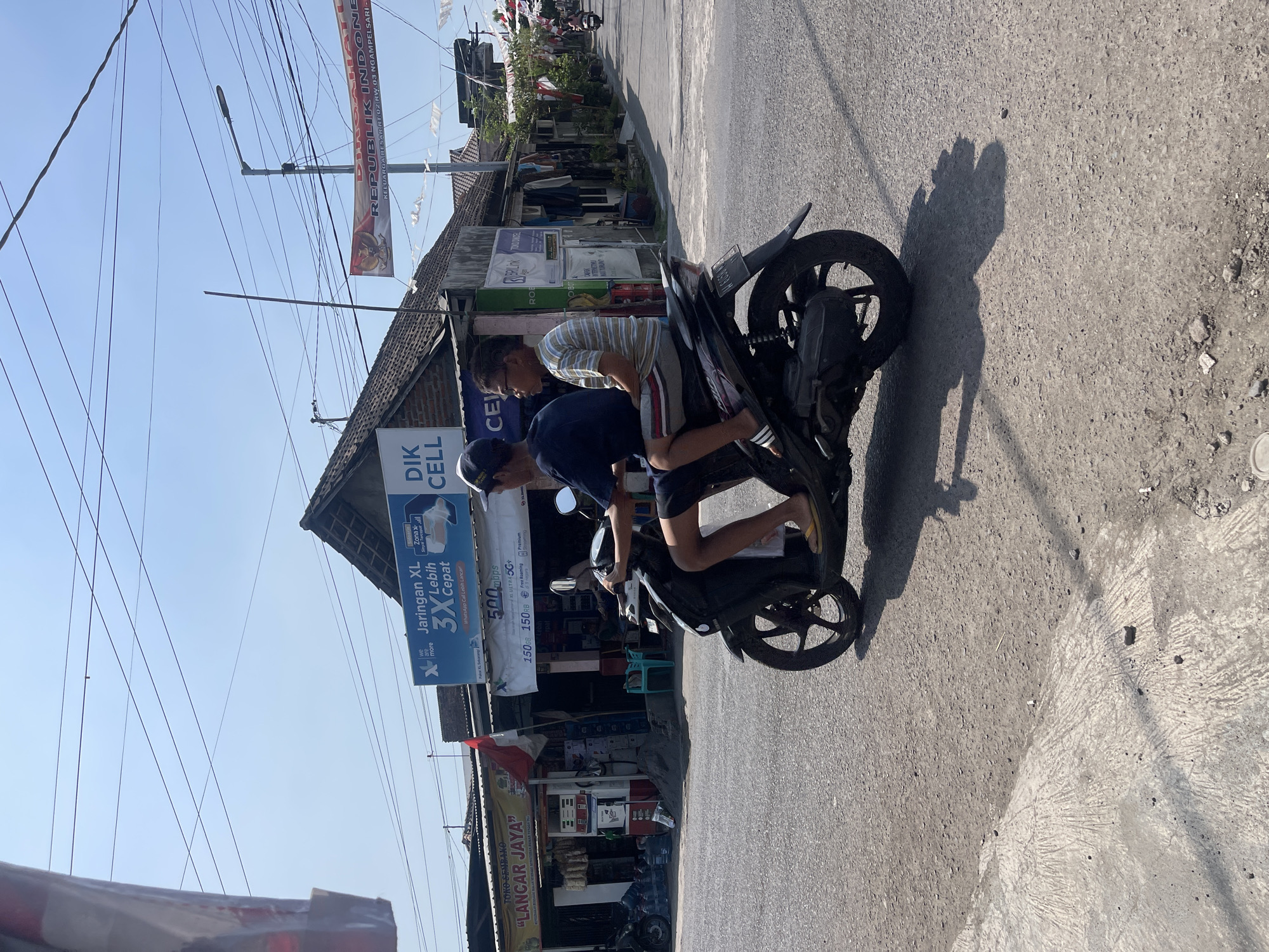 In Balonggabus, Indonesia, two individuals, an adult male and a juvenile male, are riding on a black Yamaha motorcycle. The adult male is operating the motorcycle, wearing a dark blue t-shirt and dark shorts. The juvenile male, wearing a striped t-shirt and shorts, is the passenger. The motorcycle is traveling on a paved road. In the background, a row of buildings lines the street, including a store with a sign that reads "TOKO SEMBAKO 'LANGGAR JAYA'". Another sign advertises "Jaringan XL 3X Lebih Cepat". Wires are strung across the sky above the buildings. A banner displaying the Indonesian flag and coat of arms is visible on a pole. The scene appears to be during daylight, under a clear blue sky.