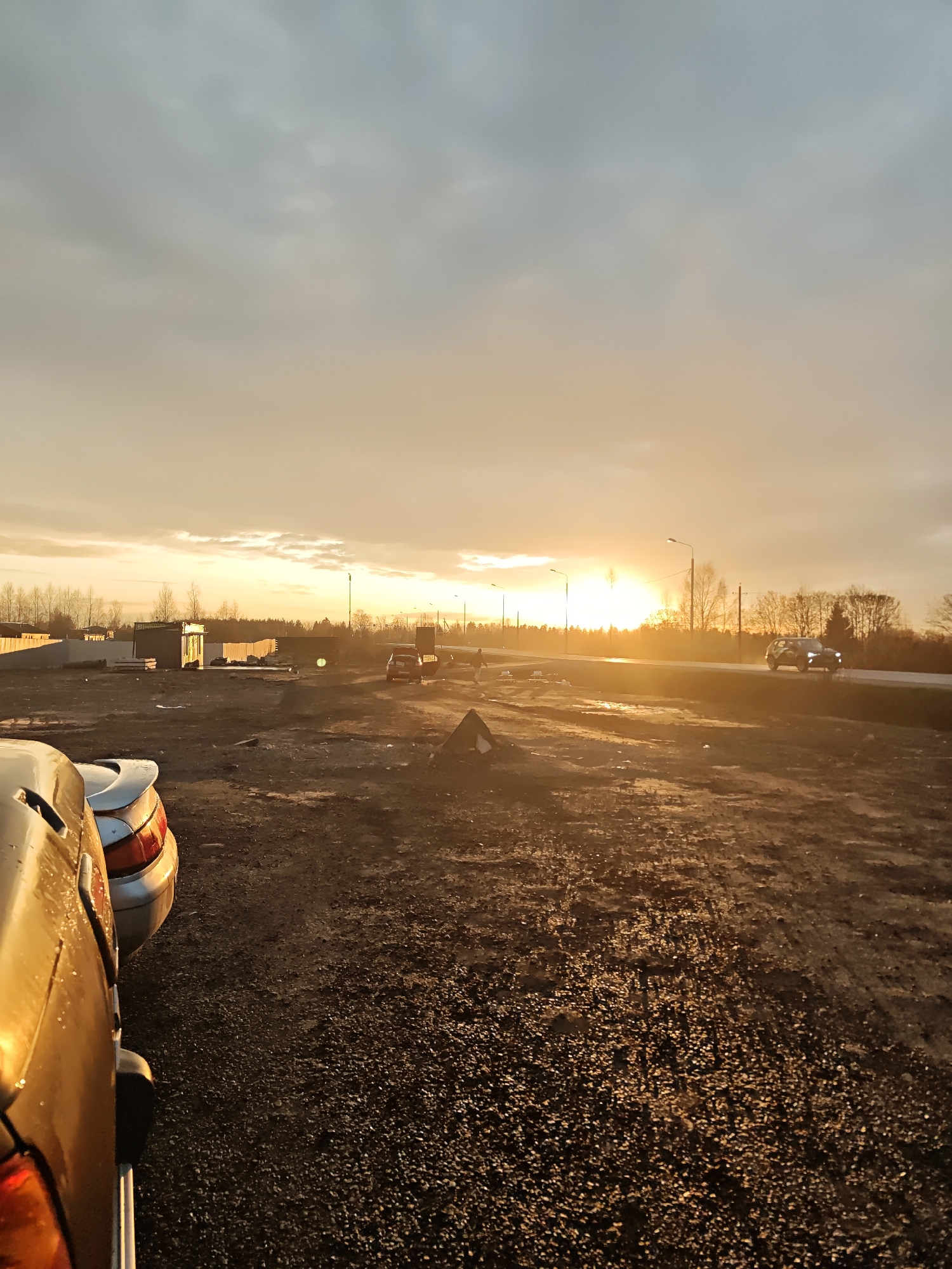 The image captures an outdoor scene at sunset in Svistukha, Russia, overlooking an open, undeveloped area adjacent to a main road. The sun, a bright orange-yellow orb, is low on the horizon, bathing the entire landscape in a warm, golden glow.
In the immediate foreground, to the left, parts of two cars are visible. The closest vehicle is metallic gold or yellow, showing its rear left side and taillight, reflecting the vivid sunlight. Behind it, a portion of a silver car's rear is also seen. The ground, a rough mixture of gravel and dirt, stretches from the foreground into the mid-ground, featuring numerous reflective patches that suggest wetness from recent precipitation.
Across the mid-ground, a paved road runs horizontally. A white car with its headlights illuminated is driving on this road, moving through the intense sunlight. Streetlights are visible along the road, silhouetted against the bright horizon. Further back, a line of bare trees, indicative of autumn, winter, or early spring, frames the scene. Several small, utilitarian buildings or sheds are scattered across the mid-ground; one features a sign with unreadable Cyrillic-like text.
The sky above presents a gradient, shifting from bright orange at the horizon to a softer, hazy grey further up, with subtle cloud formations. The overall atmosphere is quiet and dramatic, dominated by the radiant light of the setting sun.
