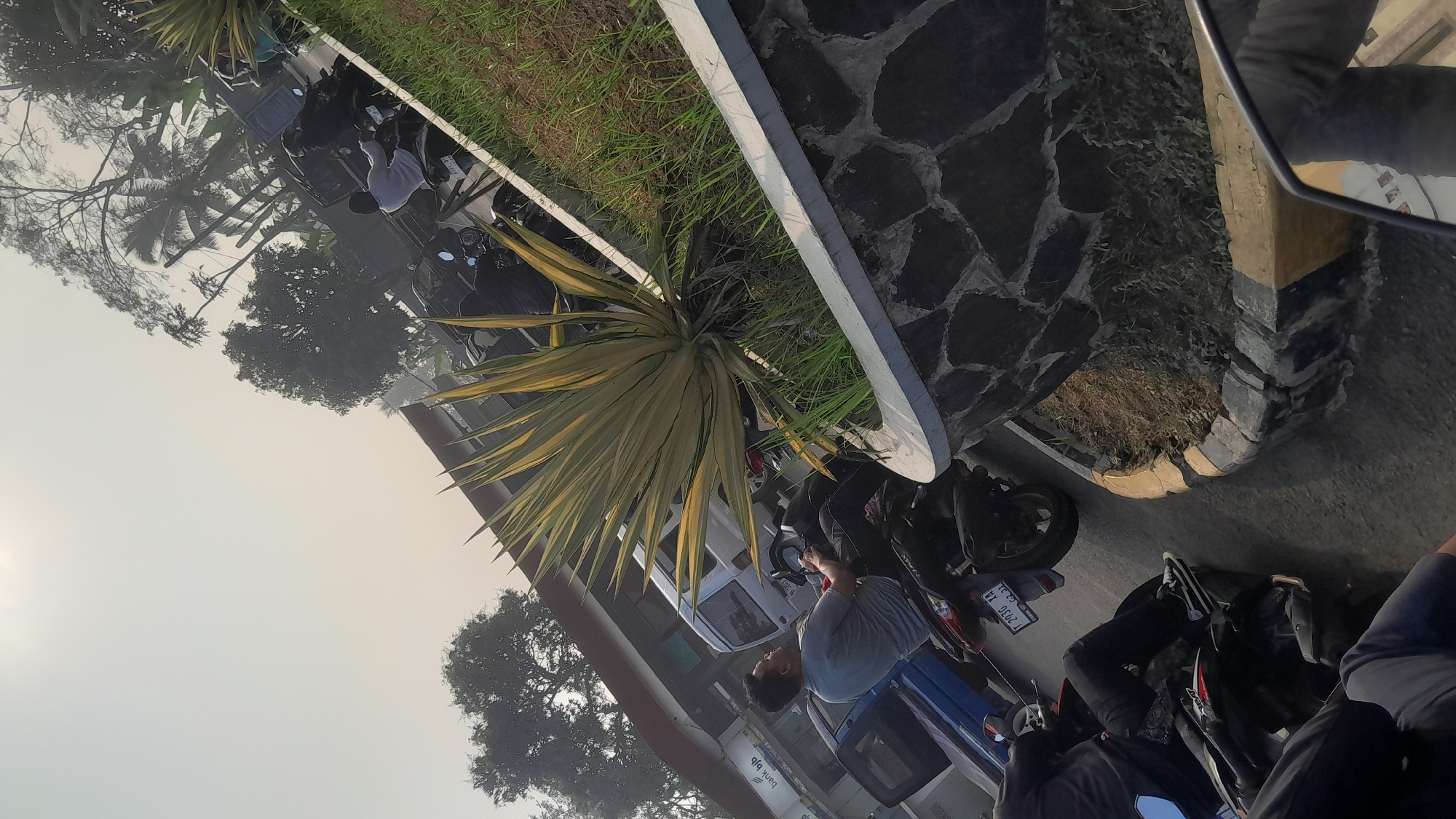The image captures a street scene in Subang, Indonesia, likely during the daytime, with a hazy sky indicating possible smog or fog. In the foreground, a person is seated on a motorcycle. The motorcycle has a license plate that reads "T 2030 XA". To the left of this motorcycle, another person on a motorcycle is partially visible, wearing black pants and white sneakers. Behind the first motorcycle, a white van is parked. Further in the background, several other motorcycles and people are visible, along with trees and buildings. A prominent agave plant is situated in a raised planter box lined with stones. The overall impression is one of a moderately busy urban environment.