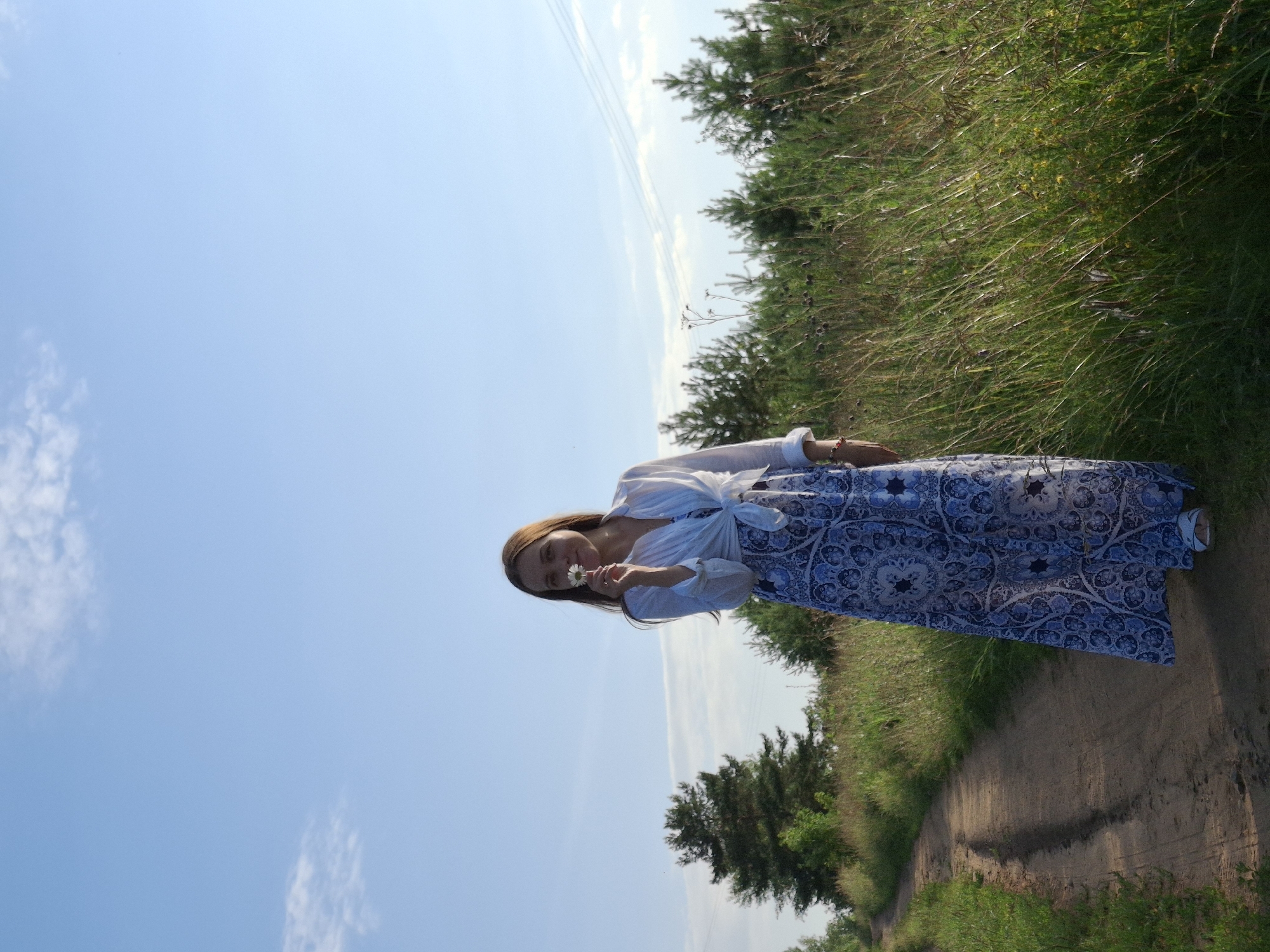 This media file shows a young woman standing on a dirt road in a grassy, rural area. She is wearing a long, blue and white patterned dress and a white shirt tied at the waist. She is holding a small white flower up to her nose, seemingly smelling it. The background consists of a clear blue sky with a few wispy clouds and a dense field of green grass and trees to her right. The scene appears to be set during the daytime, likely a warm, sunny day, given the bright light and clear sky. The woman has a gentle, perhaps contented or peaceful expression on her face. The overall setting suggests a natural, outdoor environment, possibly a park, field, or countryside road in Chaikovsky, Russia. There is no visible text in the image.