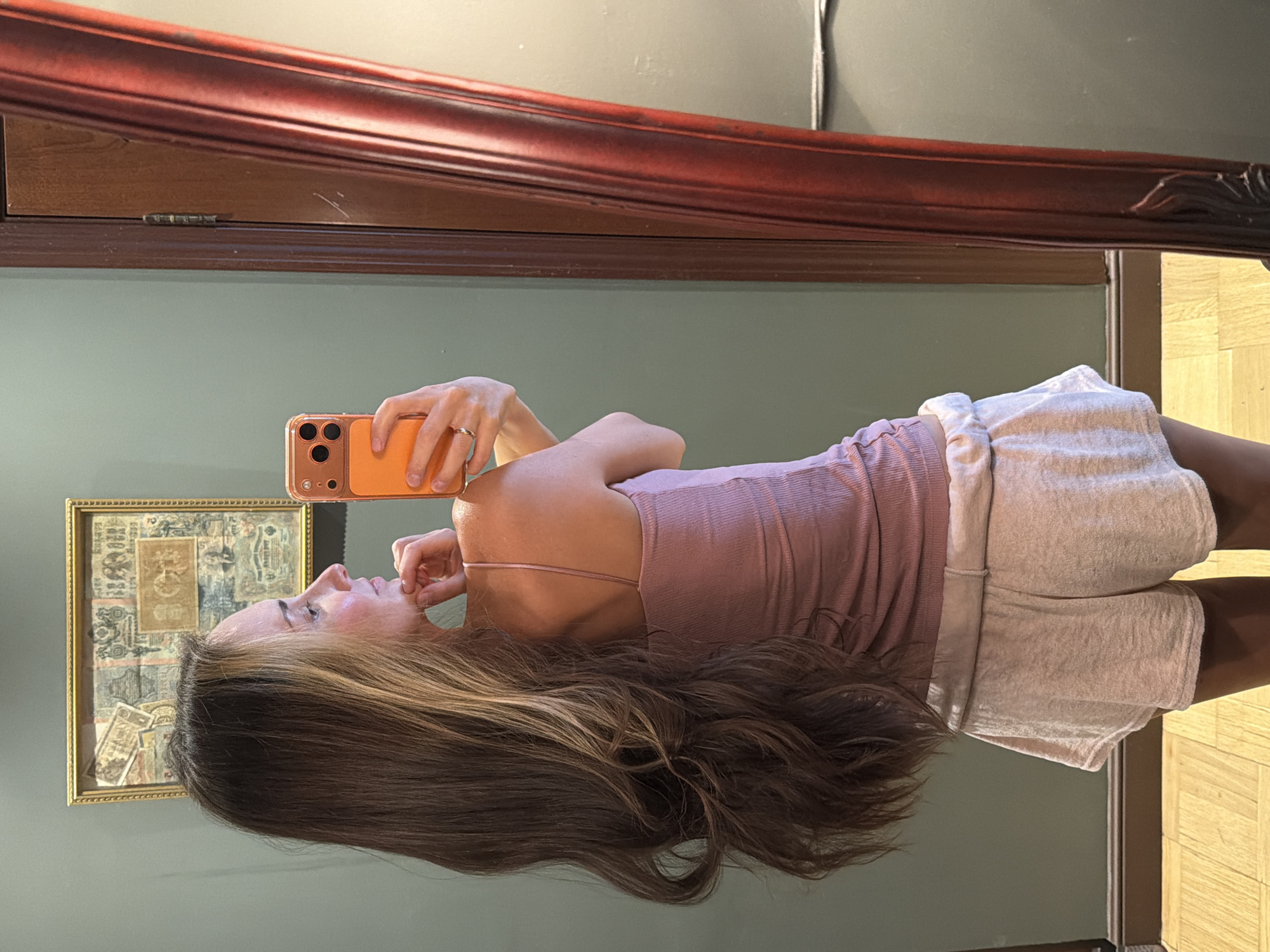 The image shows a woman taking a mirror selfie. She is standing in a room with a dark green wall and wooden floor. To the left of the woman is a framed picture hanging on the wall. To the right of the woman is a large ornate wooden mirror, its reflection showing a light-colored wall and the right side of the room. The woman is wearing a pink tank top and light-colored shorts. She has long, wavy brown hair and is holding an orange smartphone in her right hand. Her left hand is touching her chin, and she is looking to her left. The woman is wearing a ring on her left hand. There is no indication of the location being in Khimki, Russia.