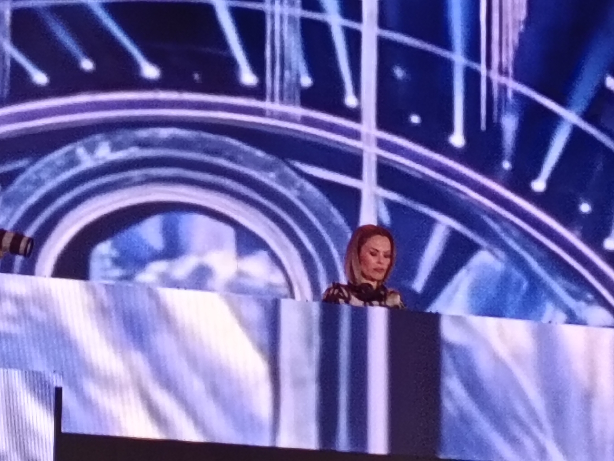 A performer, possibly a DJ, stands behind a barrier on stage in a venue that is likely an event hall, performance arena, or concert space located in San Bartolomé de Tirajana, Spain. The performer is seen from the chest up, with headphones around their neck. The backdrop features a visual display of concentric arcs with a neon or illuminated effect, punctuated by vertical streaks of light. The barrier is textured with a white and blue design. The lighting is generally blue-toned.