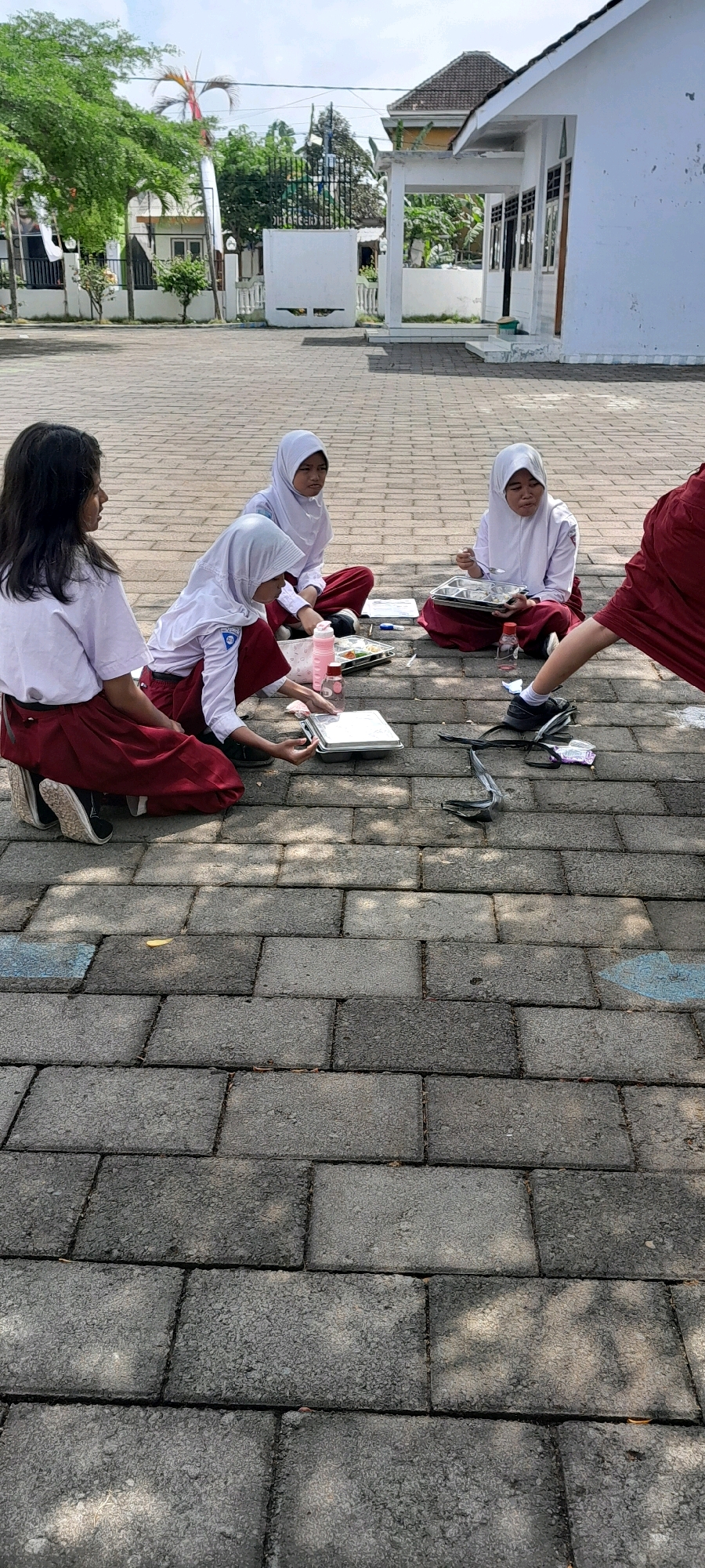 In Rogojampi, Indonesia, four young female students in white shirts and red skirts are sitting on a paved schoolyard, appearing to be eating lunch or engaged in a classroom activity outdoors. One student, facing left, has dark, shoulder-length hair and is looking away from the camera. Two other students are wearing white hijabs and are seated facing each other, with one holding a utensil and a tray of food. The fourth student, partially visible from the back, wears a red uniform and is reaching towards a tray on the ground. Visible objects include food trays, water bottles, and what appear to be school books or notebooks. The environment consists of a paved area with large, rectangular tiles, a building with white walls and a dark roof in the background, and some trees and foliage.