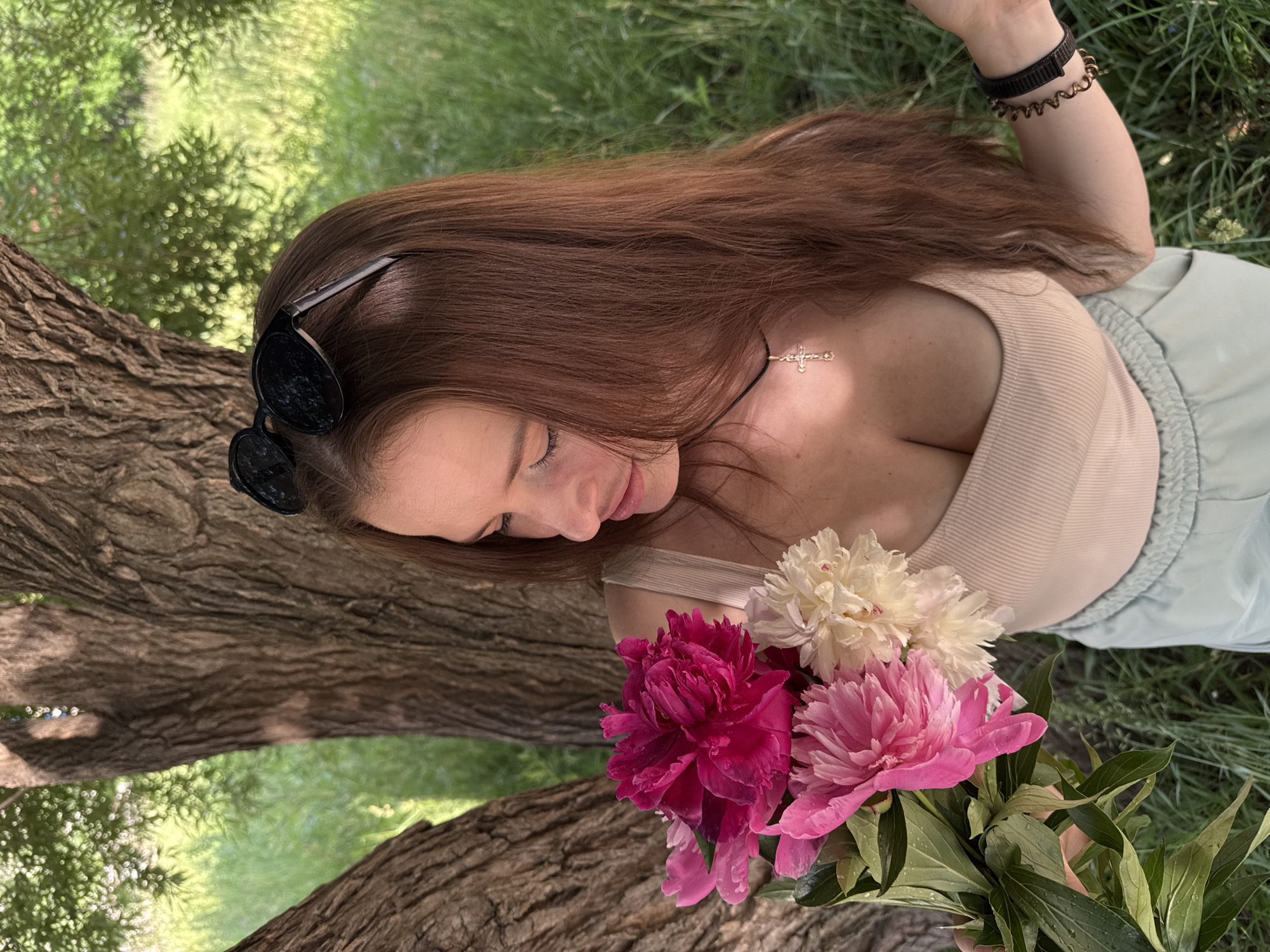 A young woman with long brown hair and a cross necklace is holding a bouquet of pink and white peonies. She is wearing a beige tank top and light blue shorts. She is sitting on grass next to a large tree. The sun is shining through the leaves of the tree, creating dappled light on the scene. The woman is looking down at the flowers, and her sunglasses are resting on her head. The background is a blur of green grass and trees. The image appears to be taken in a park or natural setting.