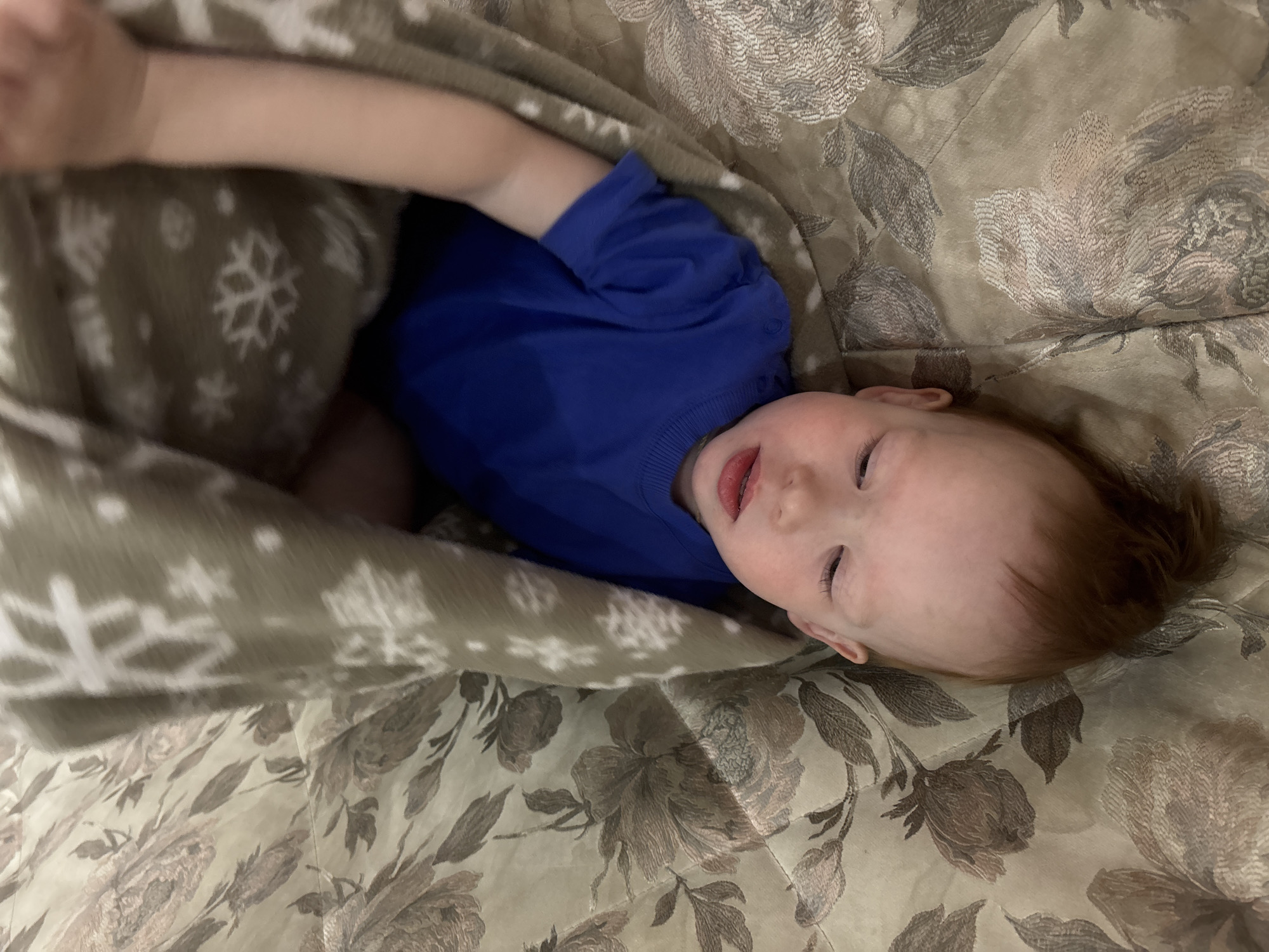 An overhead shot depicts a young child lying on their back, partially concealed by a patterned blanket, indoors in the city of Ферма, Russia. The child's head is positioned towards the bottom of the frame, with eyes closed or nearly closed, reddish-brown hair, and slightly parted lips. They are wearing a long-sleeved blue top. A light brown blanket featuring white snowflake patterns covers the upper torso and arms. The child rests on a surface, likely a bed or sofa, upholstered with a fabric exhibiting a repetitive floral pattern in shades of beige, brown, and cream.