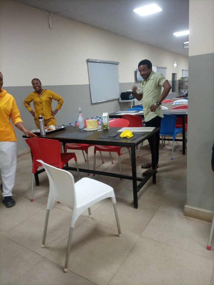 A brightly lit room features several people, tables, and chairs. Two women stand to the left, one wearing yellow and white clothing, the other wearing yellow. A man in a green shirt and black pants stands next to a table, gesturing. A whiteboard hangs on the wall in the background. The tables have red, white, and blue chairs around them. Food and drinks are arranged on the tables.