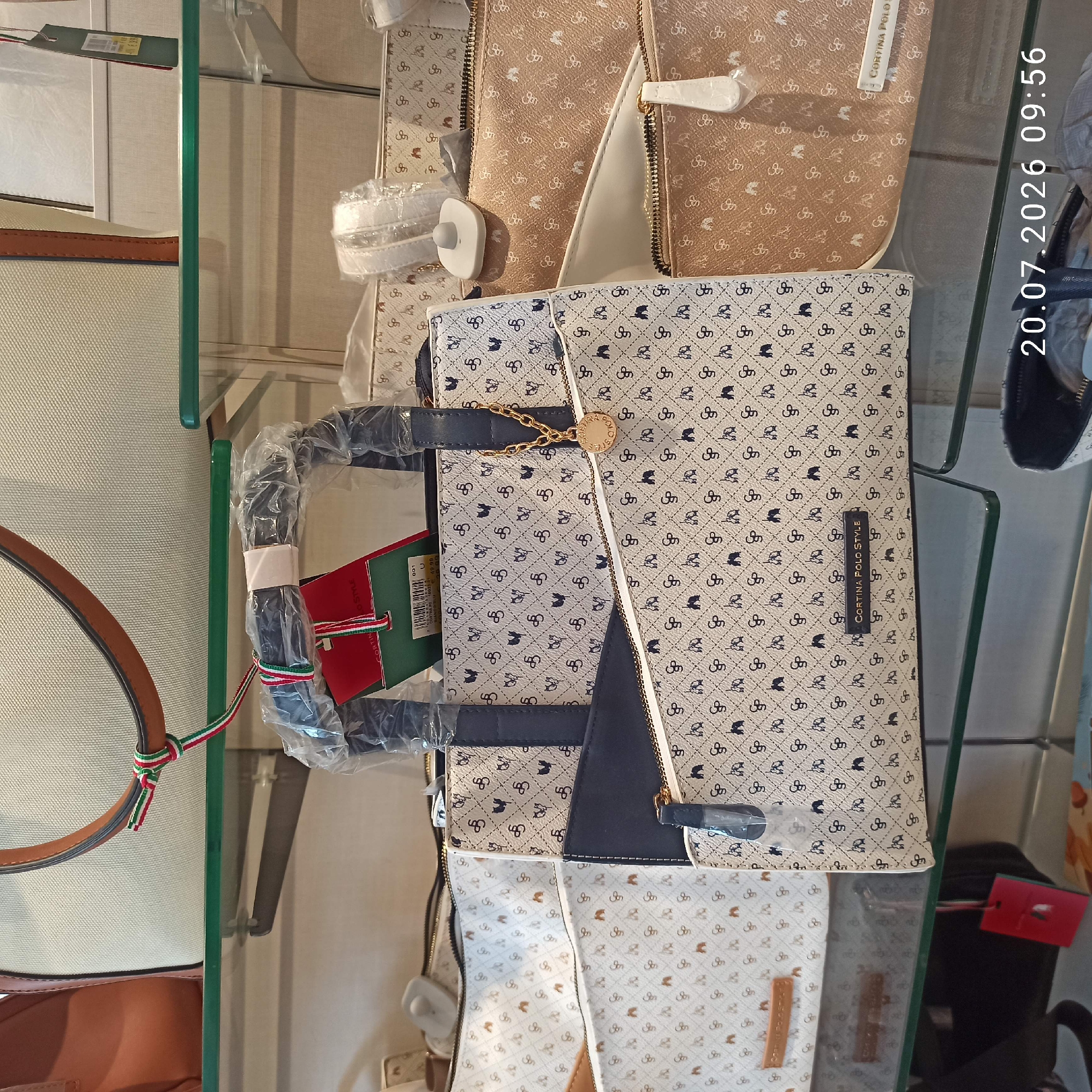 The image displays a retail environment, possibly a boutique or department store, showcasing handbags on display shelves. The focus is on a collection of handbags with a repeating pattern of intertwined "CP" logos and small animal figures, rendered in a dark color against a lighter background. The bags vary in size and style, with some featuring zippered compartments and decorative gold-tone hardware. One bag prominently displays a tag with "CORTINA POLO STYLE" printed on it, along with other product information. Other bags in the background exhibit similar patterns in different colorways, including a tan version. Security tags are visible on some of the items. The overall impression is one of merchandise presentation in a commercial setting. The timestamp overlay in the lower right corner of the image reads "20.07.2026 09:56". There is no information in the image to indicate the location in the city of Bari, Italy.