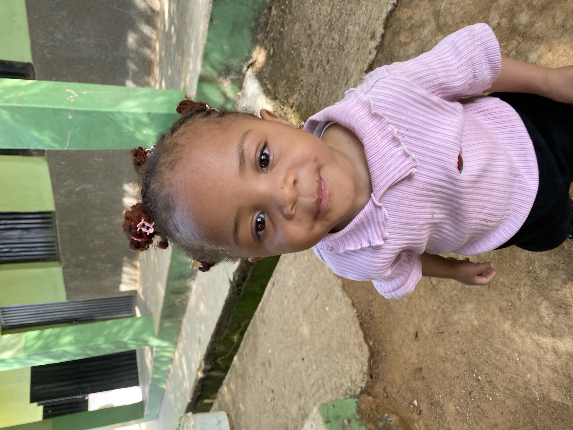 A young child is viewed from above, the camera angle looking down at them. The child has brown skin, brown eyes, and dark hair styled with several small buns adorned with pink and white hair clips. They wear a pink, ribbed shirt with a small strawberry design near the collar and black pants. The background includes a section of a green painted wall with what appear to be metal vents or openings. The ground is a mix of concrete and dirt. This scene is located in the city of Gura Top, Nigeria.