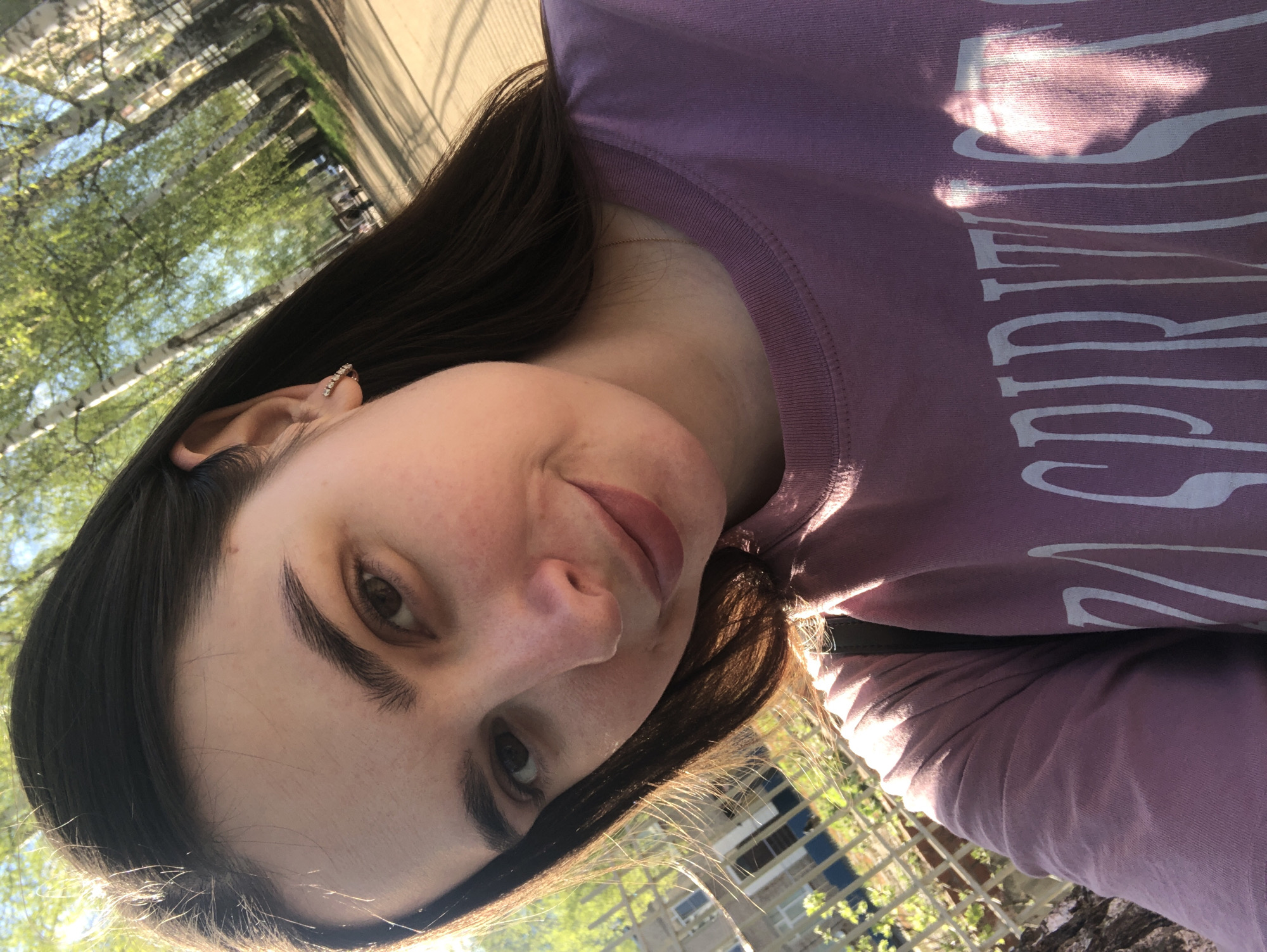 A woman with dark hair and light skin is pictured in an outdoor setting. She is wearing a pink t-shirt with white lettering. The background consists of trees, a building, and a fence. Based on the metadata, the location is likely the city of Chaikovsky, Russia. The woman is facing the camera, and the image appears to be taken at an angle.