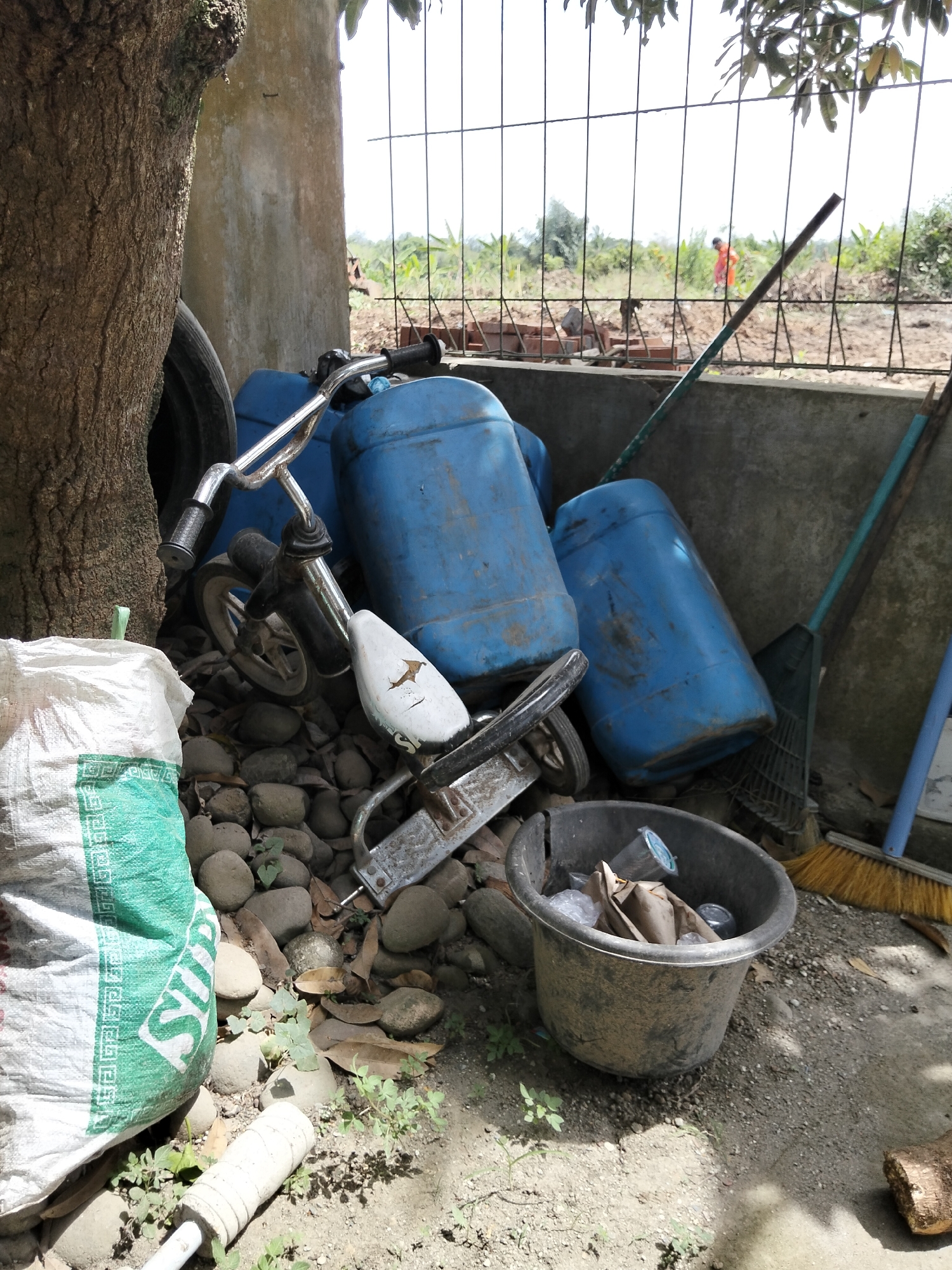 The image depicts an outdoor scene in Payagaboh, Indonesia, characterized by clutter and discarded items. In the foreground, a large white and green woven bag is leaning against a tree trunk. Nearby, a collection of rounded stones is scattered, interspersed with dried leaves and small green plants. A child's bicycle, appearing weathered, is partially visible, with its handlebars and front wheel prominent. Behind the bicycle, several large blue plastic barrels are stacked. To the right, a black plastic bucket contains various pieces of trash, including plastic cups and paper. Two brooms, one with a green handle and bristles and another with a yellow bristled head and a blue handle, are leaning against a concrete wall. In the background, a wire fence separates the immediate area from a rural landscape with vegetation and what appears to be a person in an orange garment walking in the distance. The overall impression is one of an unkempt outdoor storage area or yard.