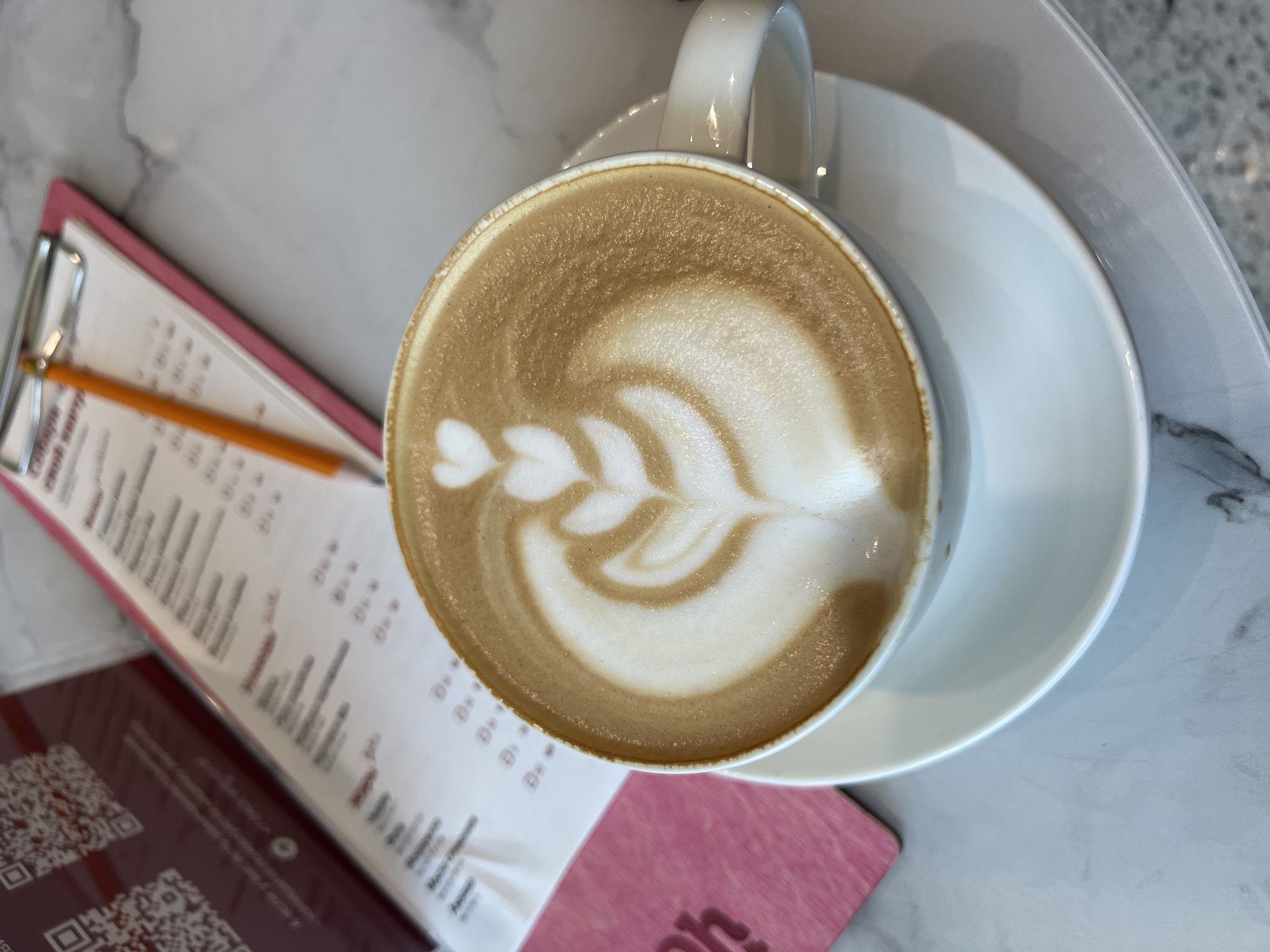 The image displays an overhead view of a light-colored table at an unspecified location in Unknown, Unknown. Centered in the foreground is a white ceramic coffee cup holding a latte adorned with elaborate white latte art, consisting of a heart shape at the top followed by a series of three progressively larger, symmetrical leaf-like patterns flowing downwards. The cup rests on a matching white ceramic saucer, with its handle visible on the right side.
In the blurred background, towards the left, a dark pink booklet or menu is partially visible, open to a page displaying two prominent QR codes and text in a non-Latin script below them. To the right of this, a light pink clipboard holds a sheet of white paper with additional text in a non-Latin script, appearing to be a menu or list with numerical or symbolic entries. An orange pencil is clipped to the top of the clipboard, resting diagonally across the paper. The table surface is light grey or white, resembling polished marble with subtle darker grey veining. No individuals are present in the scene.