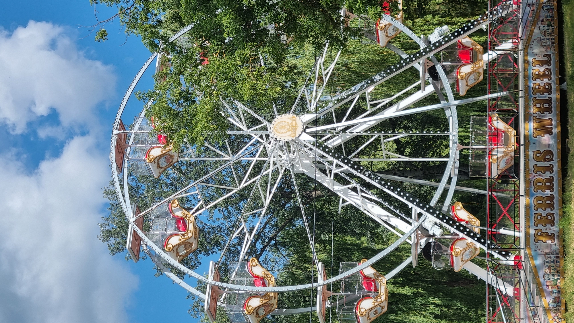 A white Ferris wheel with red and gold gondolas stands outdoors, surrounded by lush green trees under a partly cloudy blue sky. The Ferris wheel has a central medallion with the words "FERRIS WHEEL" inscribed on it. The base of the Ferris wheel is decorated with colorful imagery and the words "FERRIS WHEEL" in large, ornate lettering. The scene is set in the city of Smila, Ukraine.