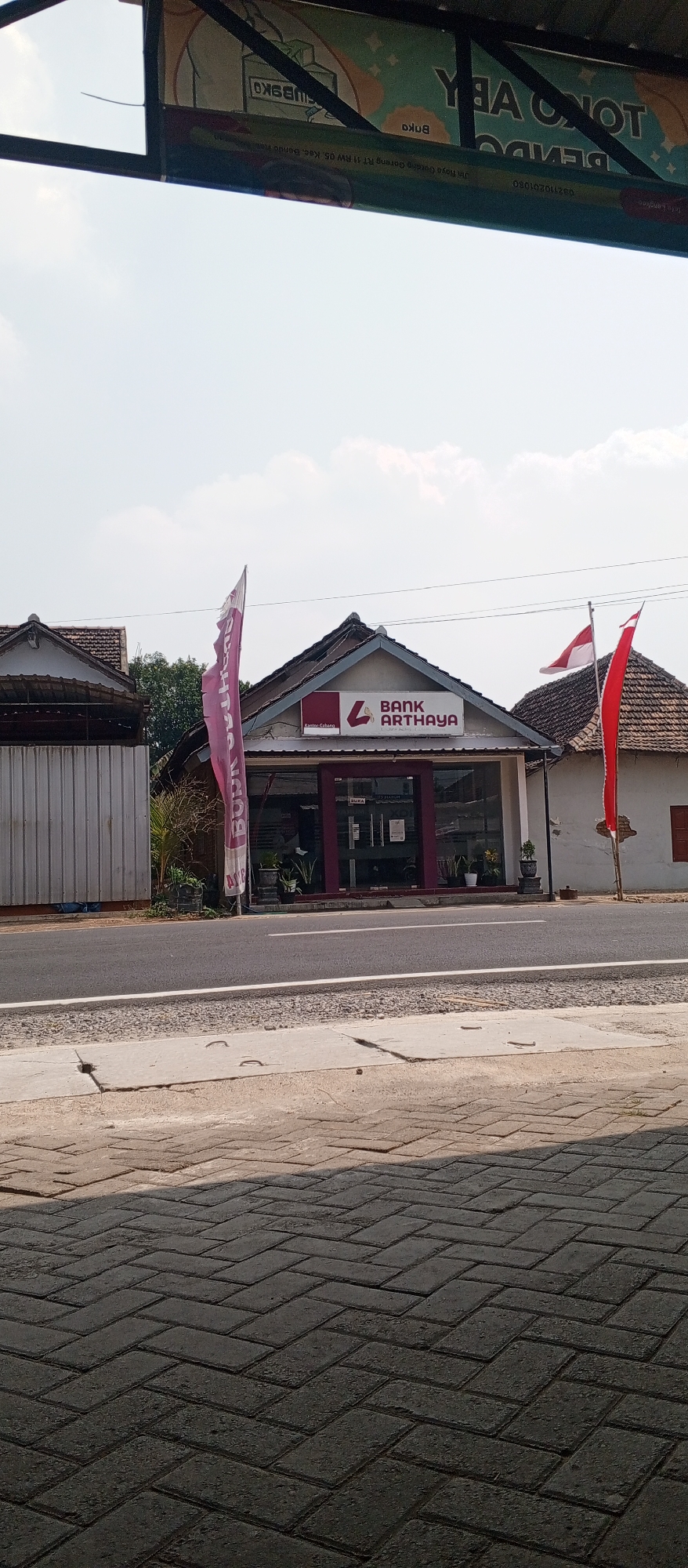 The image shows the facade of a Bank Arthaya branch in Maospati, Indonesia. The bank has a maroon-colored entrance and a sign above it displaying the bank's logo and name. Two Indonesian flags are visible, one red and white on a pole to the right of the bank, and a red flag on a pole to the left. A pink flag with text is also displayed to the left of the bank. The building has a traditional roof and appears to be situated on a street with a paved sidewalk and a road. The sky is clear with some clouds. The foreground shows a patterned brick walkway.