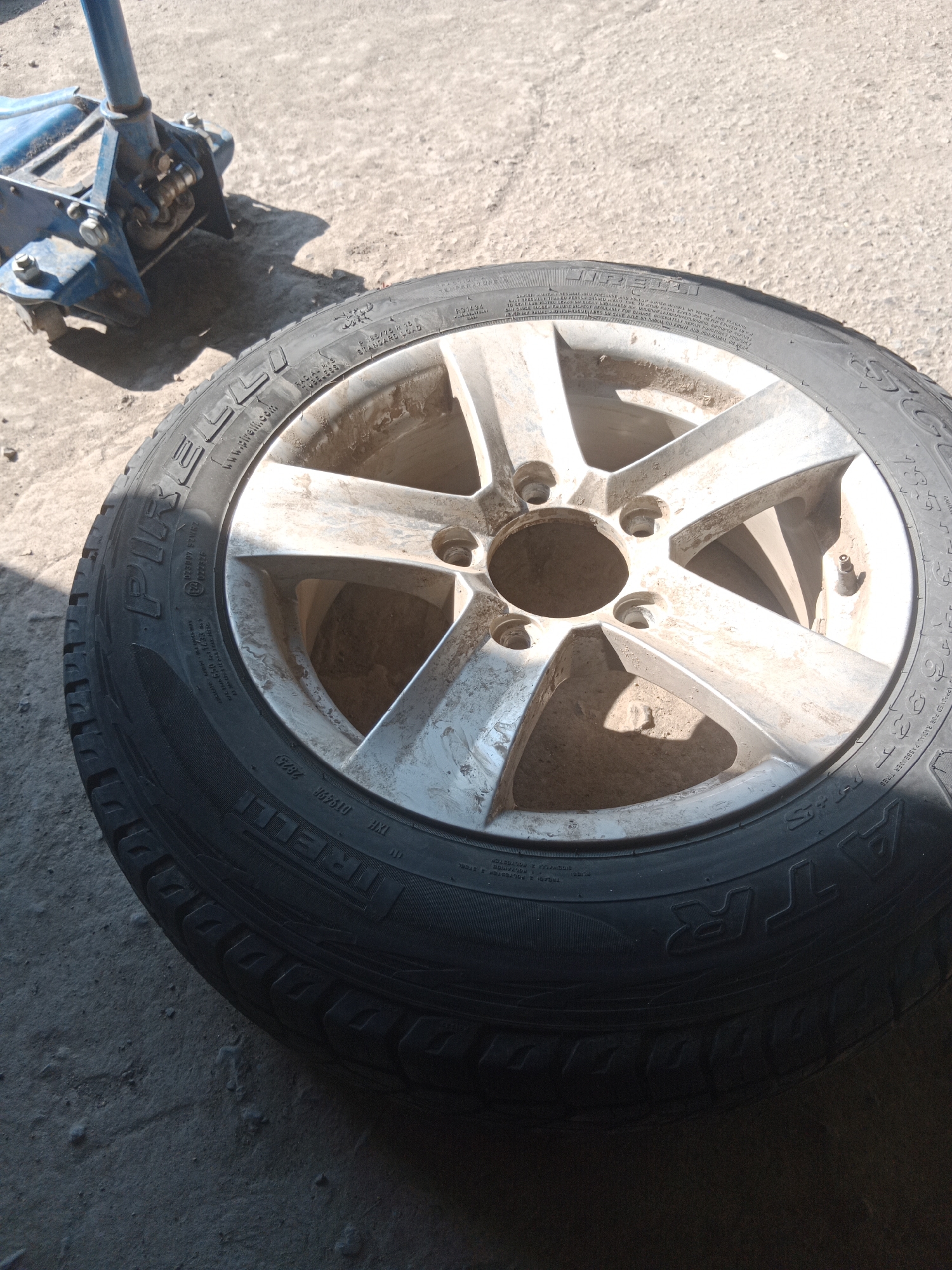 A close-up, high-angle shot shows a Pirelli all-terrain tire and a blue hydraulic car jack resting on a concrete surface. The tire, which appears to be dirty and used, is positioned in the foreground, slightly angled towards the right. The rim of the wheel is visible through the tire, showing five spokes and a silver finish. Text is visible on the sidewall of the tire, indicating brand and specifications. The car jack is situated in the background on the left side of the image. The concrete surface is textured and appears to be outdoors, with shadows cast by the objects. The scene is likely set in an outdoor urban environment, possibly a garage or workshop area.