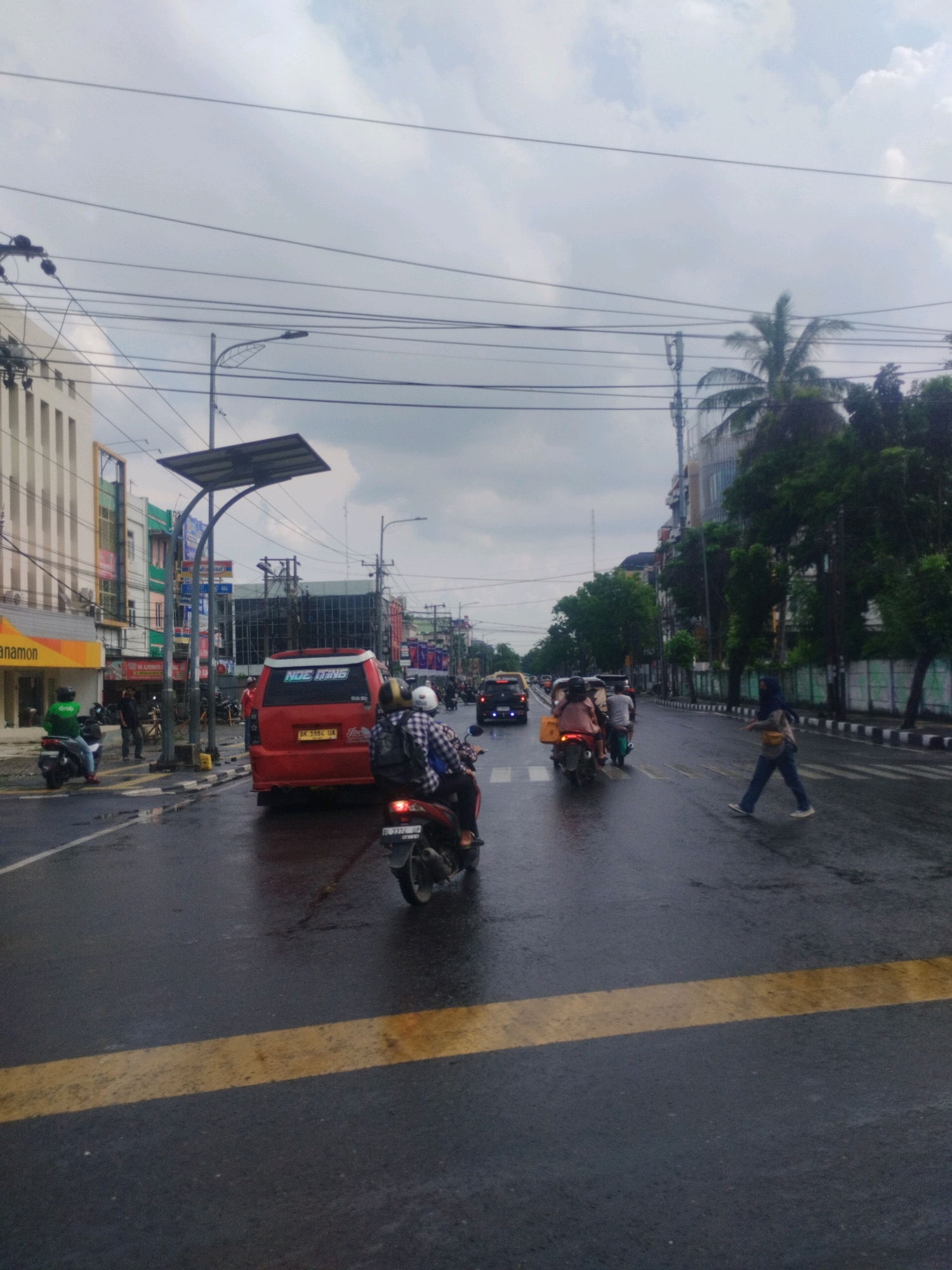 The image captures a wet city street in Medan, Indonesia, under an overcast sky. The road is slick with rain, reflecting the overcast sky and the surrounding urban environment. Several vehicles, including a red van with the text "NOE TIEMS" on its back, and multiple motorcycles, navigate the street. Pedestrians are also present; one woman in a hijab is crossing the street at a marked crosswalk, while others are visible on the sidewalks and behind vehicles. Buildings line both sides of the street, featuring a mix of commercial establishments with signage and apartment or office buildings. Overhead, numerous electrical wires crisscross the sky, characteristic of urban infrastructure. A street lamp with a solar panel attached is visible in the mid-ground, positioned over a sidewalk. The overall scene depicts a busy, active street in a tropical city during or immediately after rainfall.