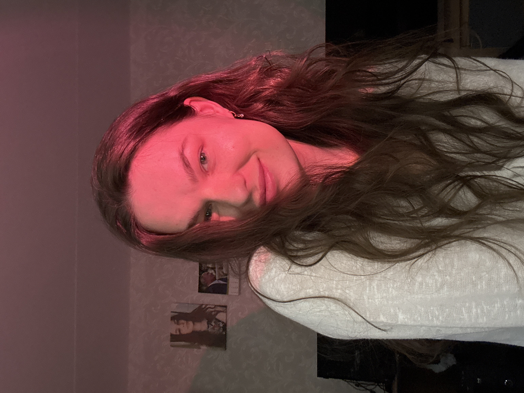 The image shows a medium shot of a young woman with long, wavy brown hair. She is wearing a white, loosely knit sweater. The lighting is predominantly red, casting a strong hue across her face and hair. She is looking towards the camera with a slight smile. In the background, on a textured wall, there are two framed pictures: one of a woman and another of a man in a suit. To the left of the woman, a black object, possibly a television or furniture, is partially visible. The overall environment appears to be an indoor residential space. The location is identified as Chaikovsky, Russia.