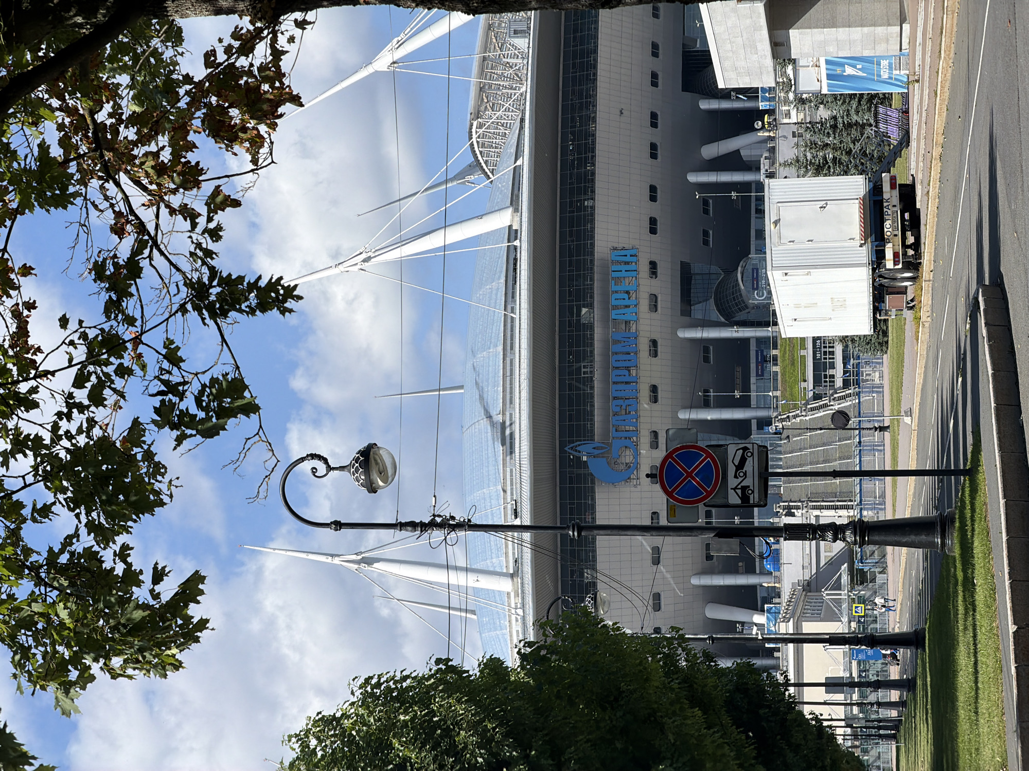 The image shows Gazprom Arena, a stadium in Saint Petersburg, Russia. In the foreground, a street is lined with trees and street lamps, with a traffic sign indicating no parking and towing. Beyond the street, the stadium's distinctive white, sail-like roof structure is visible, with large blue lettering on the facade spelling out "GAZPROM ARENA". A white portable toilet trailer is parked on the side of the road, with the word "ТУАЛЕТ" (toilet) written on it in Russian. Several people are walking on a pathway in the background. The sky is blue with scattered clouds.