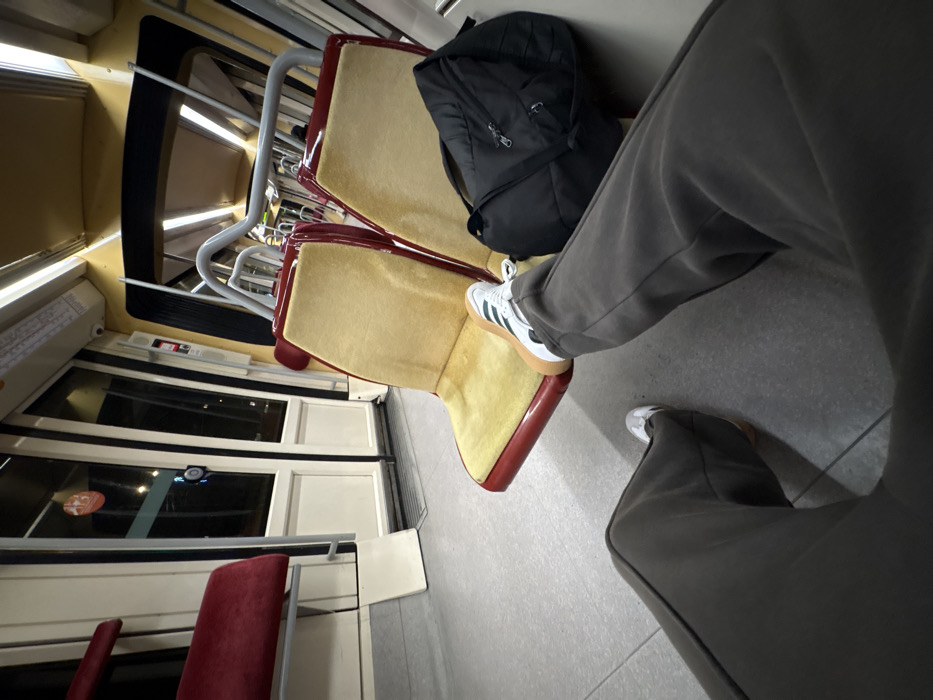 Tan and maroon train seats are occupied by a black backpack and a person wearing gray sweatpants and sneakers with white uppers and tan soles. One foot rests on the seat in front, while the other is on the floor. Windows line the left side, with a glimpse of red seating further down.