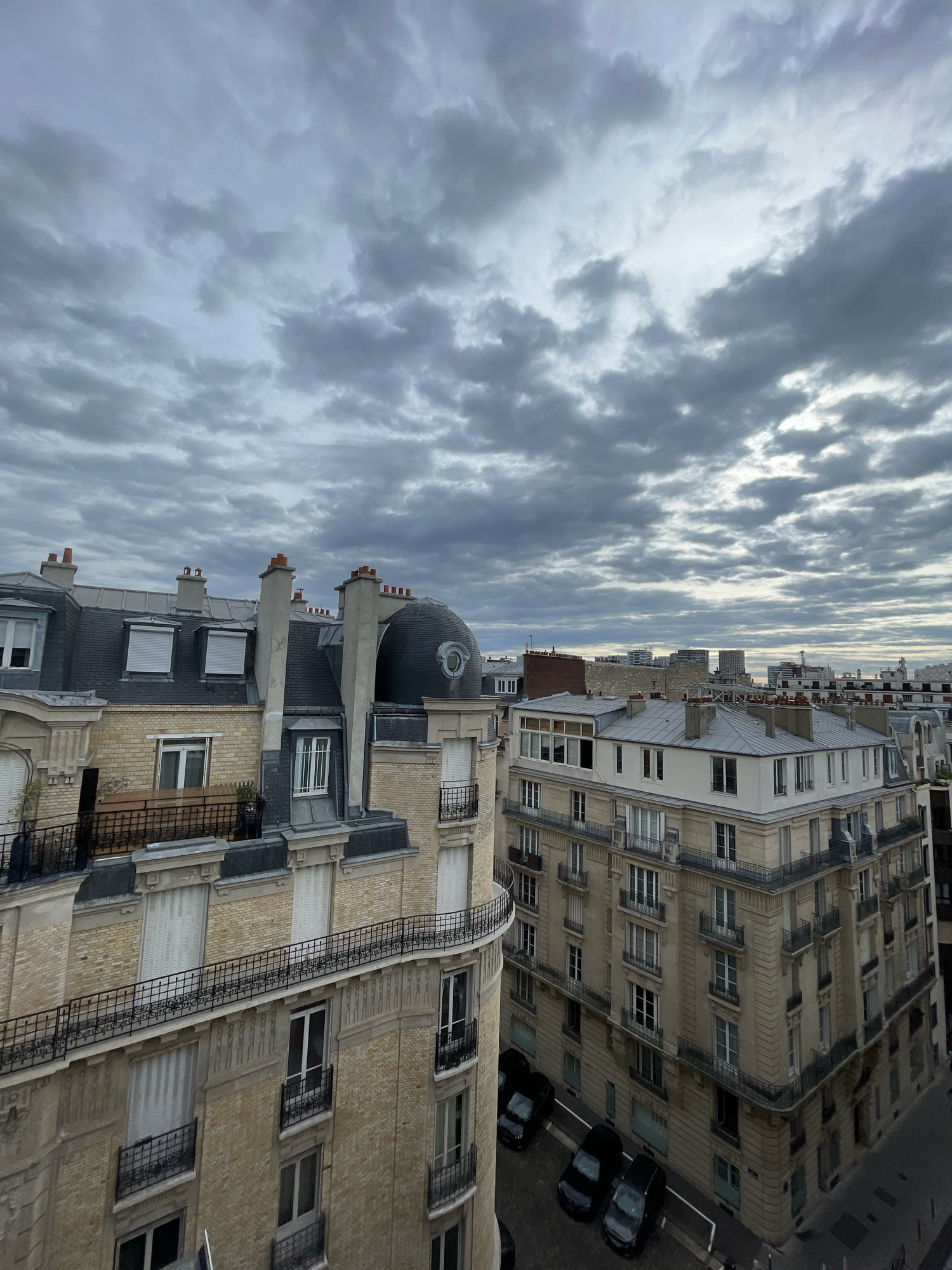 This is an aerial view of Parisian rooftops under a cloudy sky. The architecture is consistent with the Haussmannian style, characterized by cream-colored stone buildings with mansard roofs. Chimneys are prominent on the slate-tiled roofs. One building on the left features a distinctive dark dome with a circular window. Balconies with black wrought-iron railings are visible on several floors of the buildings. In the street below, several dark-colored cars are parked. The overall impression is a dense urban landscape under an overcast sky, indicative of a late autumn or winter day in Paris.