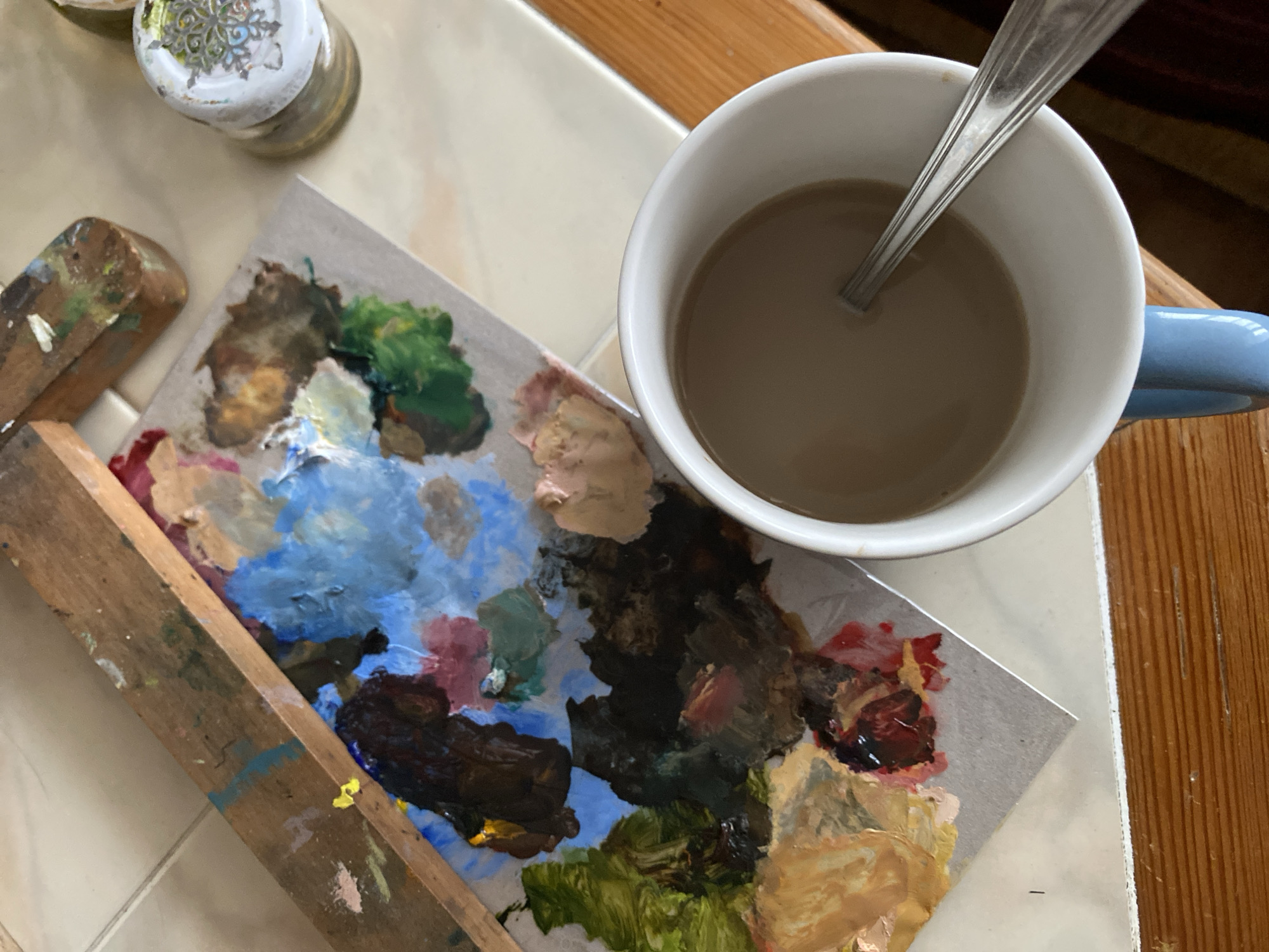 The image shows a close-up, overhead view of an art palette with various oil paint colors mixed on it, a wooden tool, a small jar with a decorated lid, and a white mug filled with coffee, containing a spoon. The palette is on a surface that appears to be tiled, with a wooden border visible in the foreground. The art supplies suggest an artist's workspace, possibly in Bílina, Czechia, though no specific landmarks or identifying features of the city are visible in the image. The lighting is natural, casting soft shadows. The scene is static and appears to be a moment captured during a break from artistic activity.