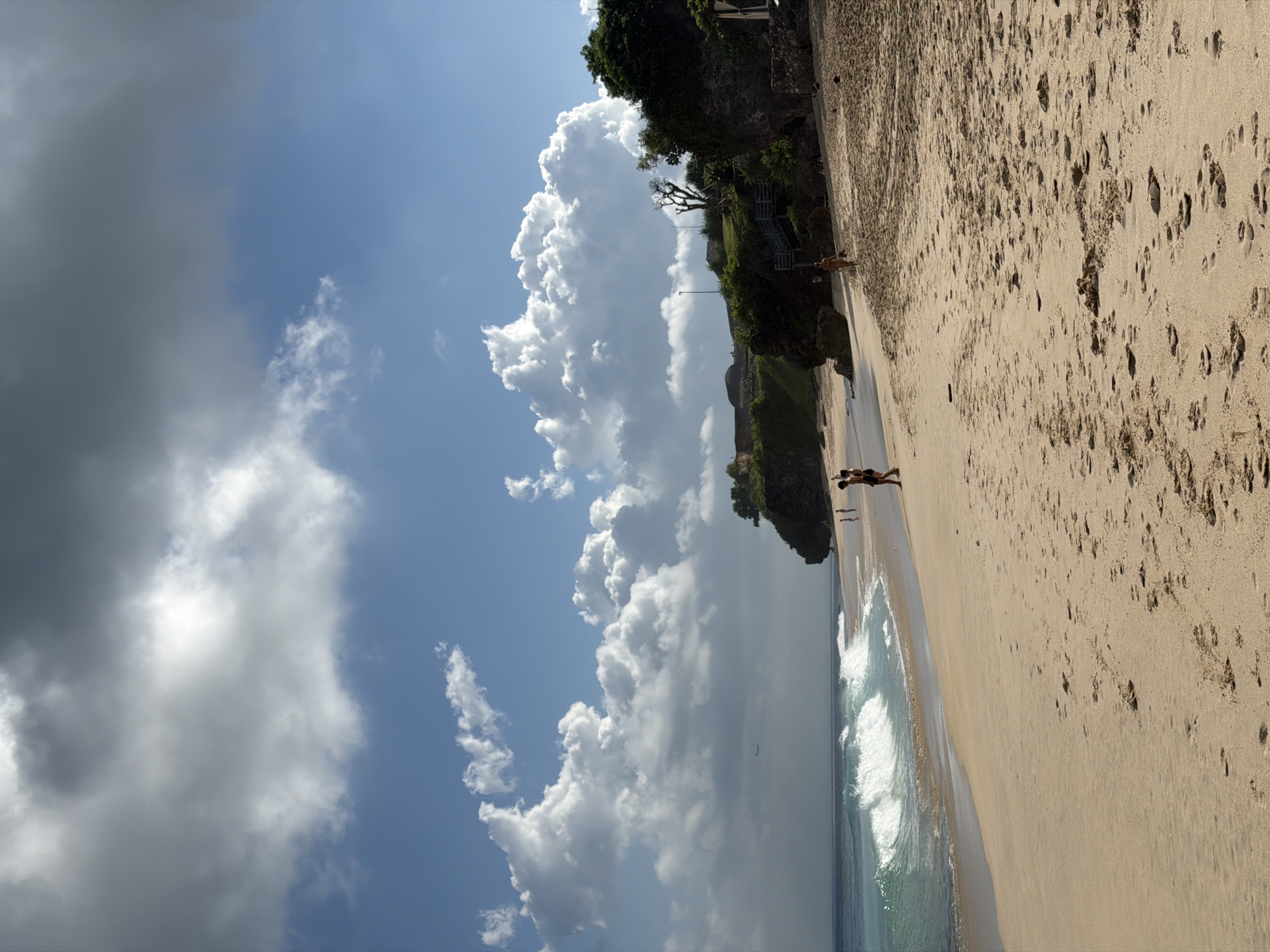 The image depicts a beach in Kuta Selatan, Indonesia. The sandy beach extends from the bottom right, marked with numerous footprints, towards a shoreline where waves are breaking. Along the top, a cliff face supports greenery. Two figures are lying on the beach near the cliff. Above, the sky is partly covered with heavy clouds.
