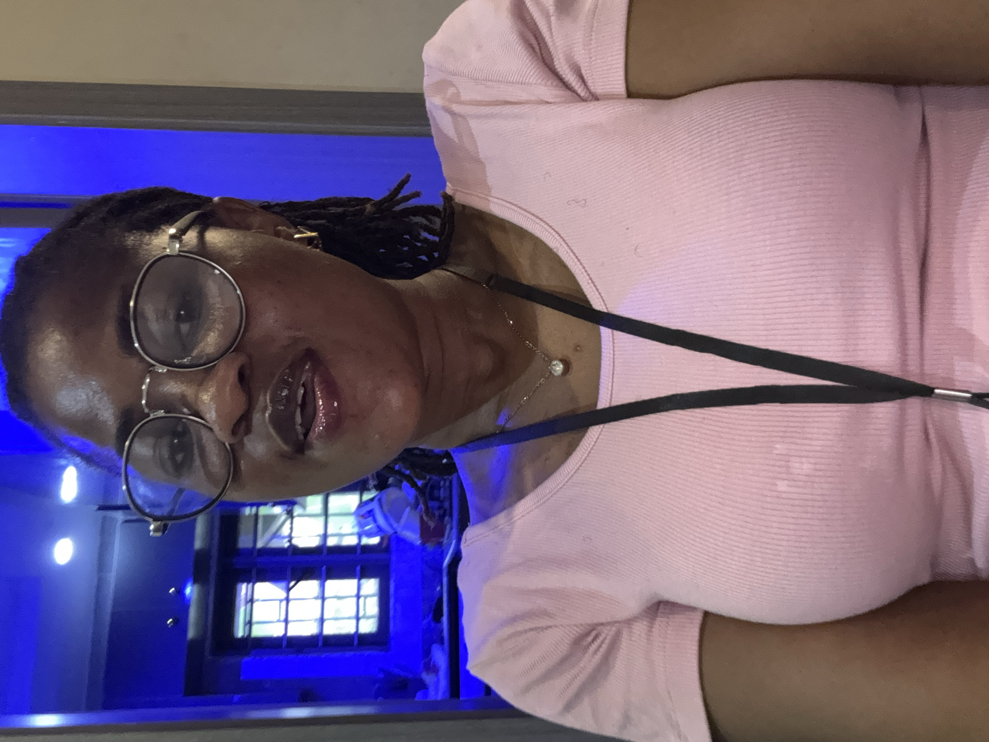 A woman with braided hair and glasses is pictured from the chest up. She is wearing a pink, ribbed t-shirt with a black lanyard around her neck. Her mouth is slightly open and she appears to be speaking. The background is out of focus and shows a bright blue light and what appears to be a window with grids. The image is taken indoors, possibly in a public building or a space with decorative lighting. The overall impression is a candid shot, possibly a selfie, given the angle and close-up framing of the subject. The scene is brightly lit, with the blue light creating a vibrant and somewhat abstract backdrop.