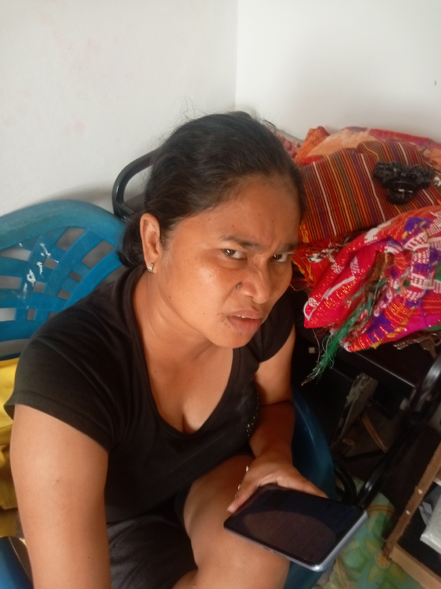 A woman with dark hair and brown skin is sitting in a blue chair, looking at the camera with a grimace. She is wearing a black t-shirt and dark shorts. A smartphone is in her right hand, resting on her lap. In the background, there are colorful fabrics and a white wall. The scene appears to be indoors, likely in a residential setting, and could be located in Batam, Indonesia, given the context.