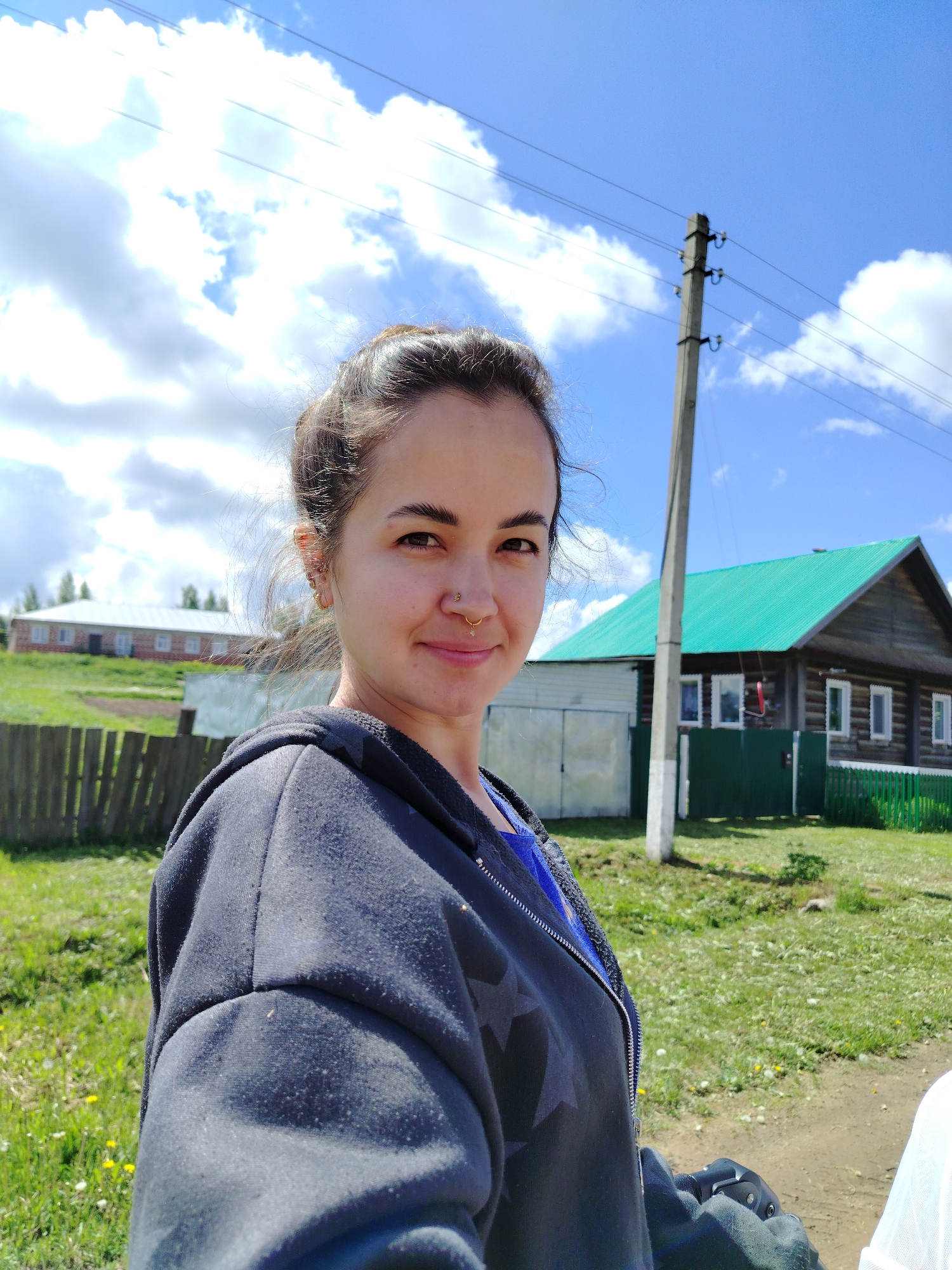 A woman takes a selfie in Гожан, Russia on a sunny day. She has dark hair pulled back and wears a black hoodie with star patterns over a blue shirt. The woman has a nose ring and multiple earrings. Behind her is a rural scene with a weathered wooden fence, a green-roofed wooden house, and a utility pole. Further back is a long, brick building. The sky is blue with puffy white clouds. The ground is green and grassy.