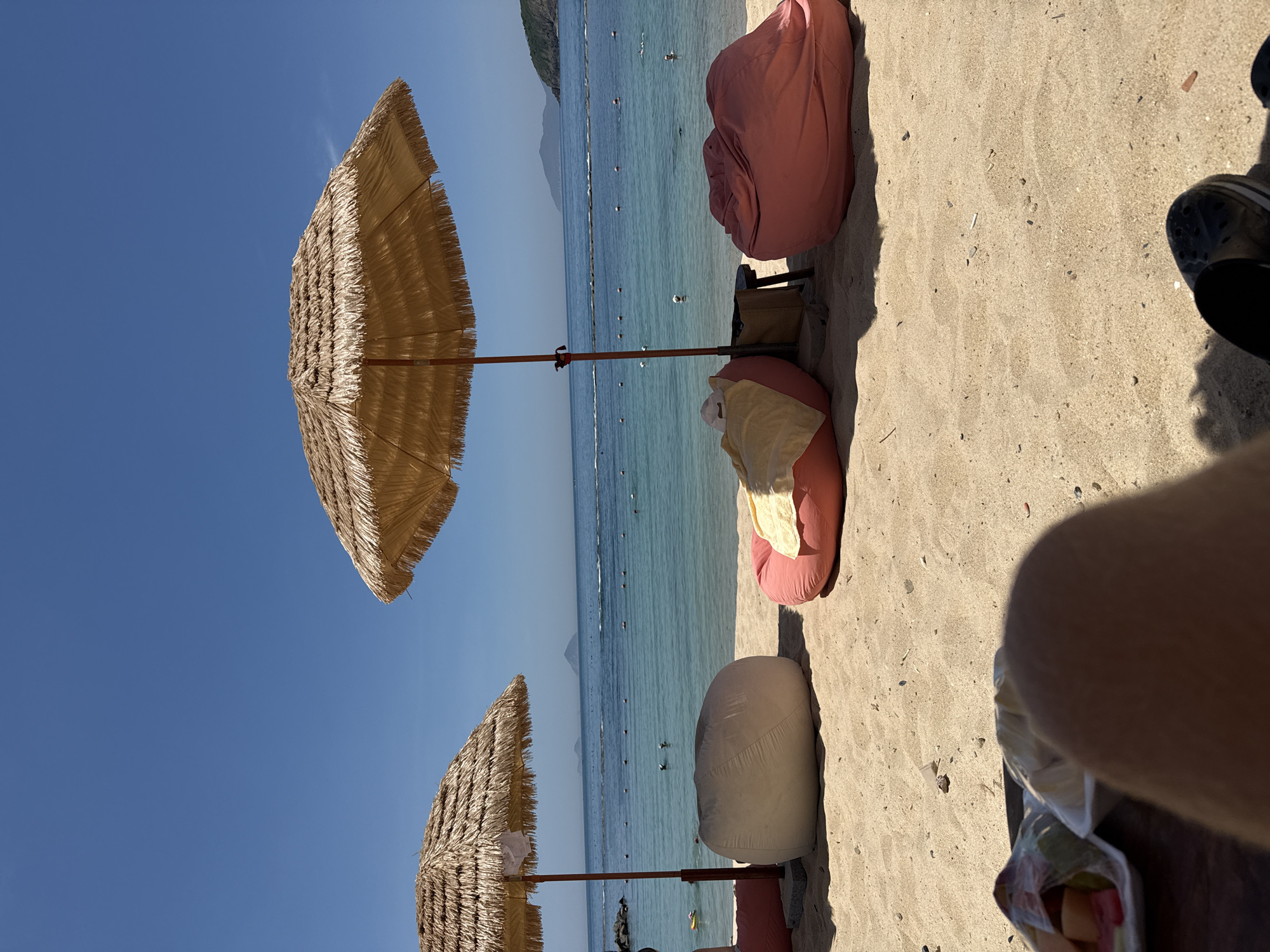 This media file captures a beach scene with two thatched umbrellas providing shade on a sandy shore. In the foreground, beanbag chairs in pink and beige are arranged on the sand. The calm, turquoise water stretches out to the horizon, dotted with buoys and a few distant swimmers. A mountainous coastline is visible in the background under a clear, bright blue sky. The lighting suggests it is a sunny day, likely mid-morning or afternoon. The overall atmosphere appears relaxed and peaceful, characteristic of a beach vacation. The image is oriented vertically, with the sky at the top and the beach sand in the foreground.