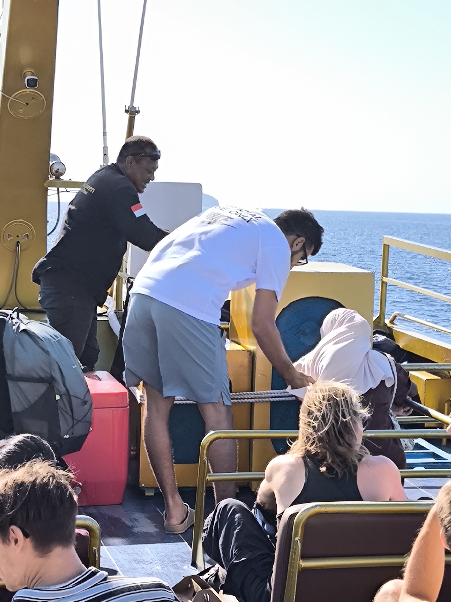 The photograph captures a sunny scene on a boat deck, likely in a coastal region of Indonesia, possibly near Unknown. People are seen interacting and seated, with the ocean visible in the background.
An Indonesian man stands on the deck, wearing a black jacket adorned with the Indonesian flag. Next to him, another man in a white T-shirt and light gray shorts assists a woman who is seated and wearing a hijab. A woman with blonde, wind-blown hair sits nearby in a dark tank top.
Bags and a red cooler sit on the deck near the passengers. Seating is arranged around the deck with the ocean clearly in the background. The boat's metallic structure is yellow in color. A small security camera is affixed to the side of the metallic structure.